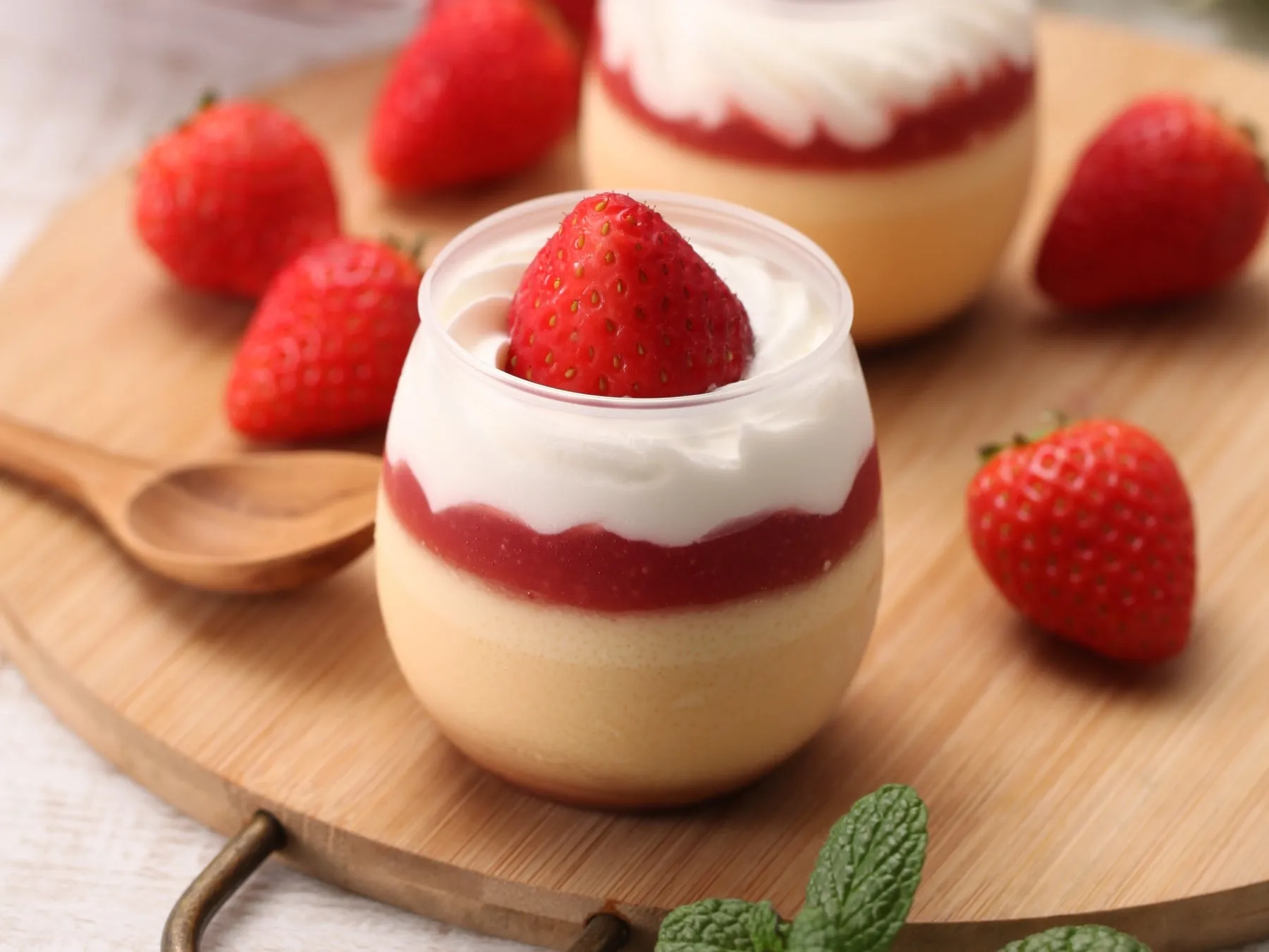 いちごのデザートがいくつか乗った木製のボードのクローズアップです。デザートは、透明なカップに入った層状のデザートで、クリーム、イチゴソース、クリーム、トッピングとしてイチゴを添えています。周囲には新鮮なイチゴが散らばっており、ミントの葉も添えられています。