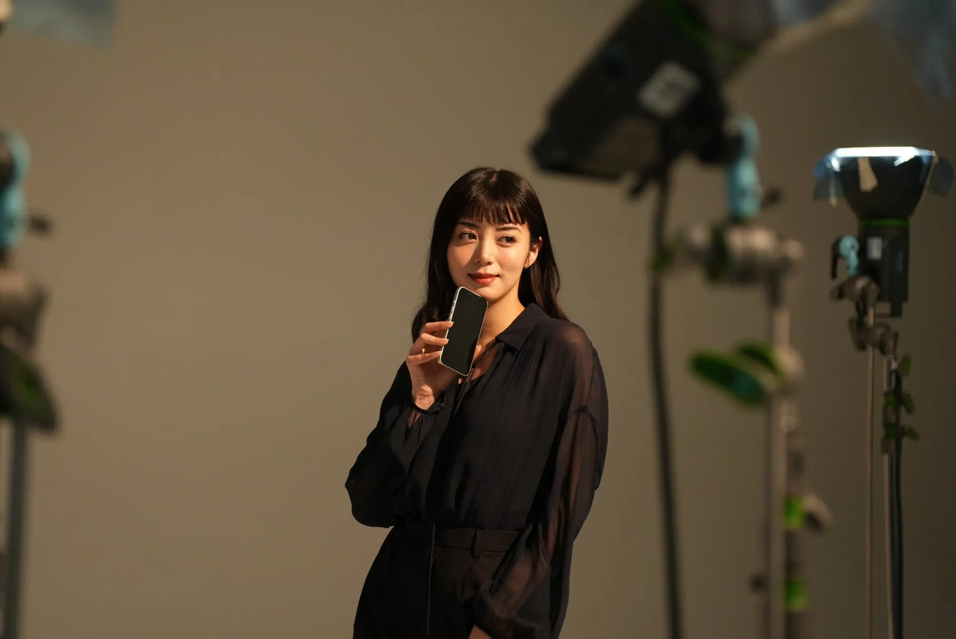 スタジオでスマートフォンを持ち、カメラを見ている女性。背景には照明器具が見える。