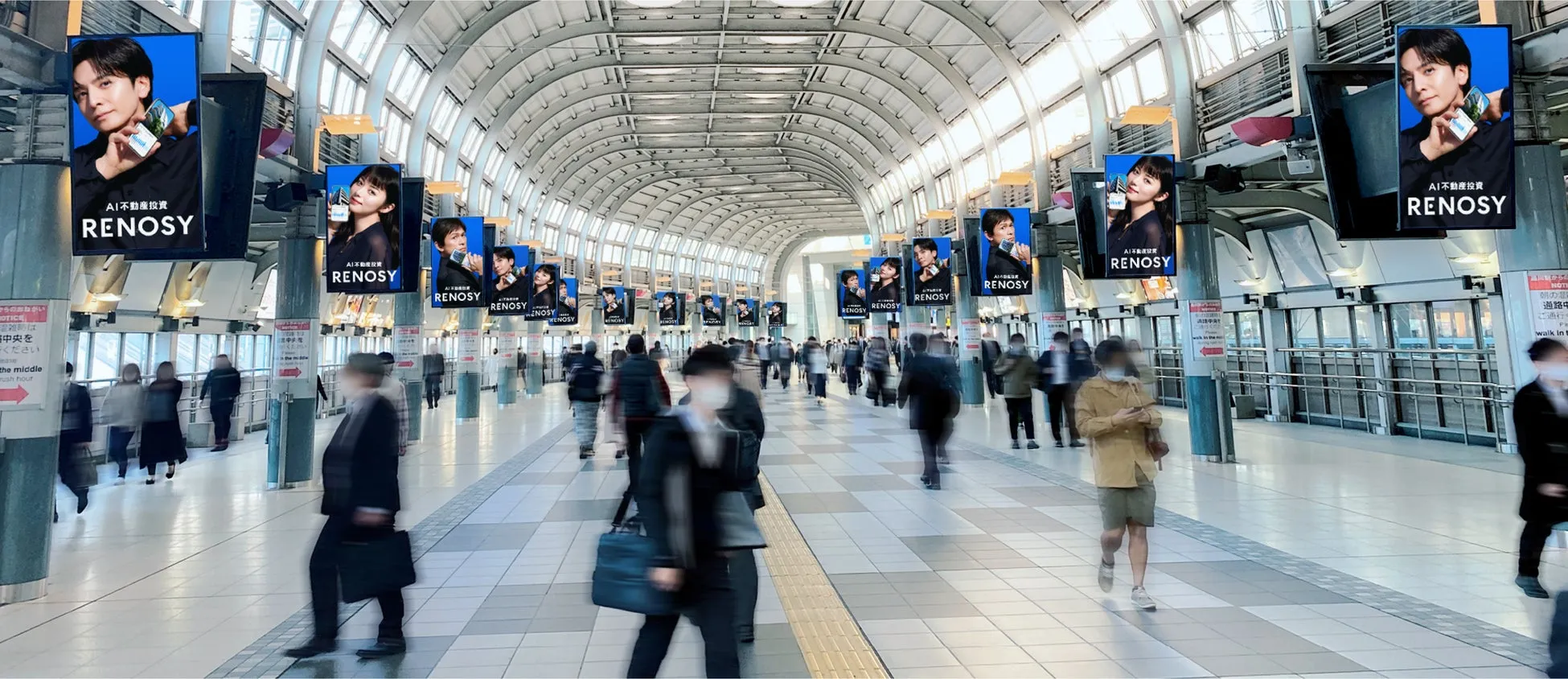 駅構内の広告看板。RENOSYの広告が多数掲示されている。多くの人が行き交い、活気のある様子。