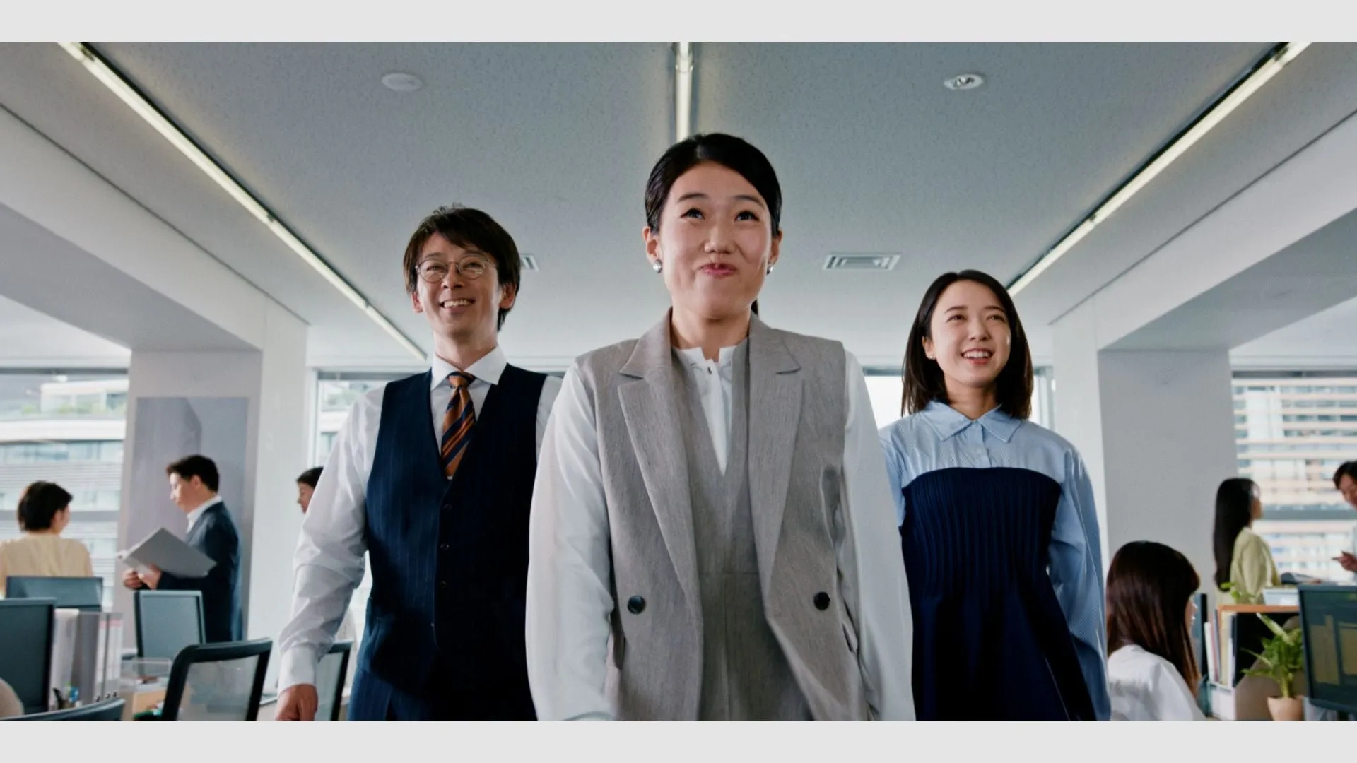 オフィスで笑顔で歩く3人のビジネスパーソン。他の従業員も写っている。