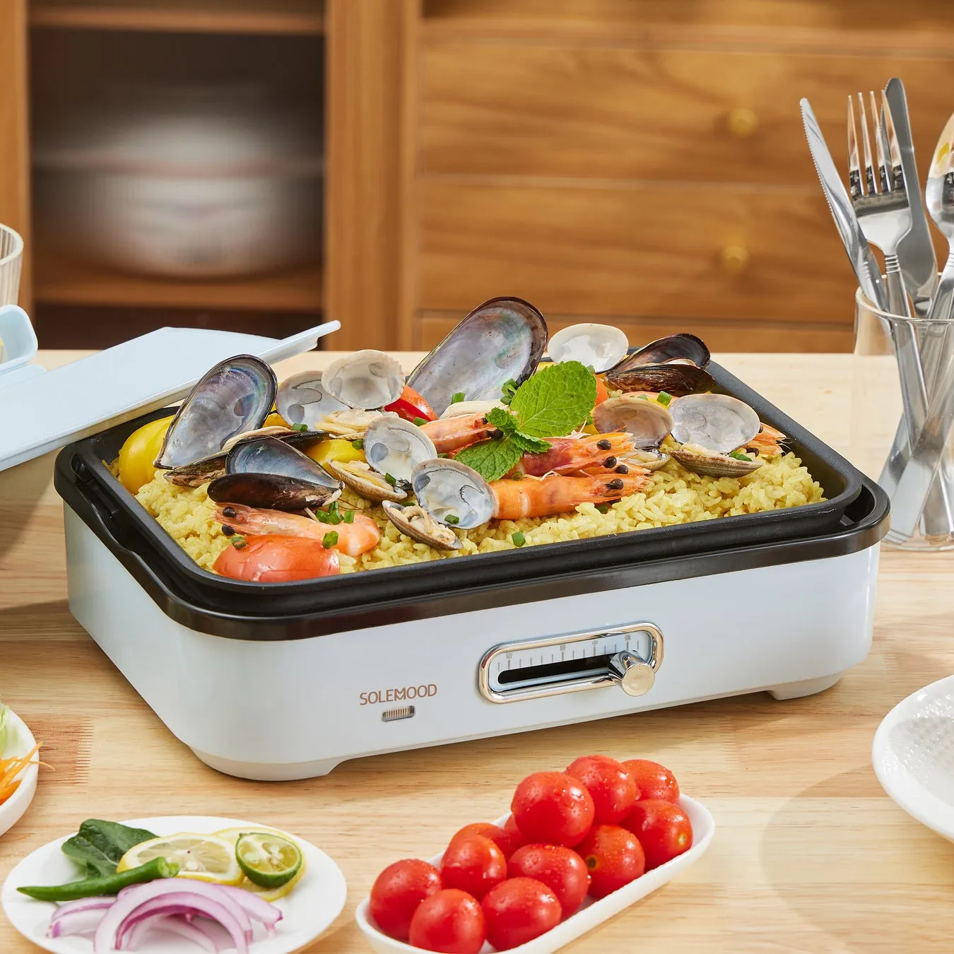 テーブルの上に、シーフードパエリアが載った電気グリルと、トマトや野菜が添えられた料理の写真です。