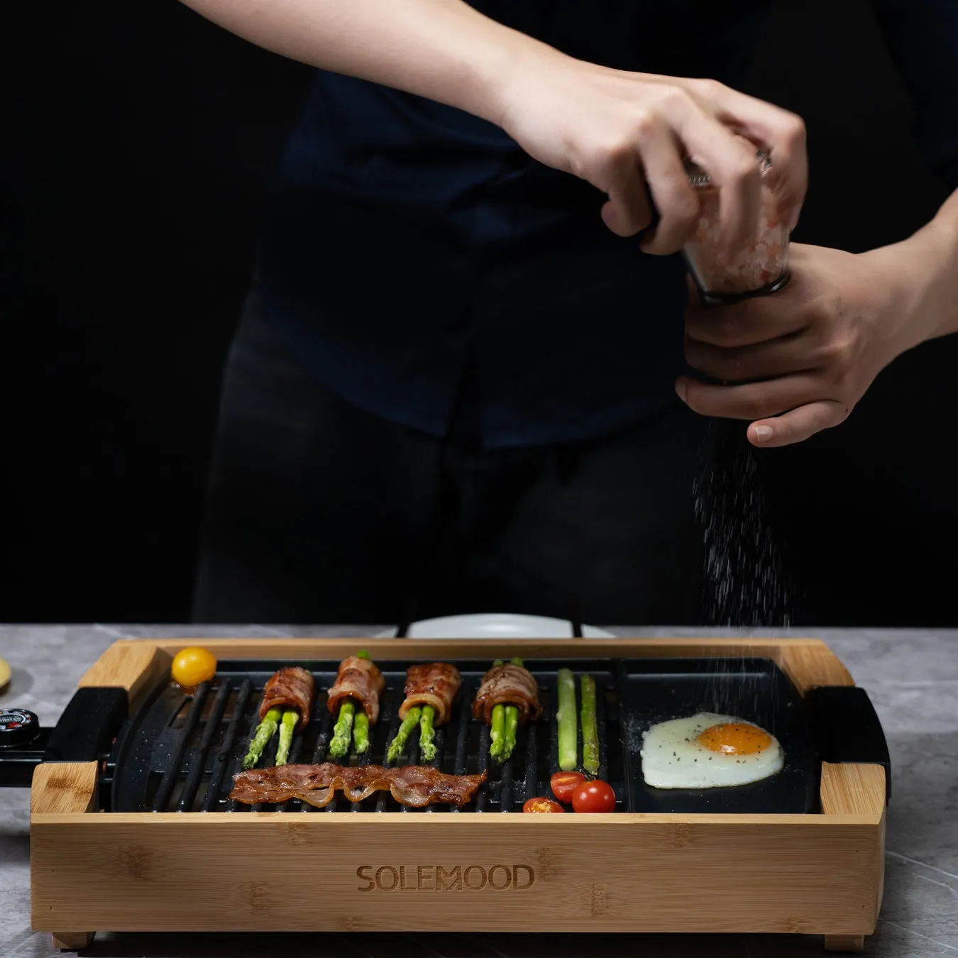 グリルで調理された料理に塩を振る人の画像です。アスパラガスをベーコンで包んだもの、トマト、目玉焼き、グリルされたベーコンが写っています。グリルは木製のフレームで、SOLEMOODというブランド名が書かれています。