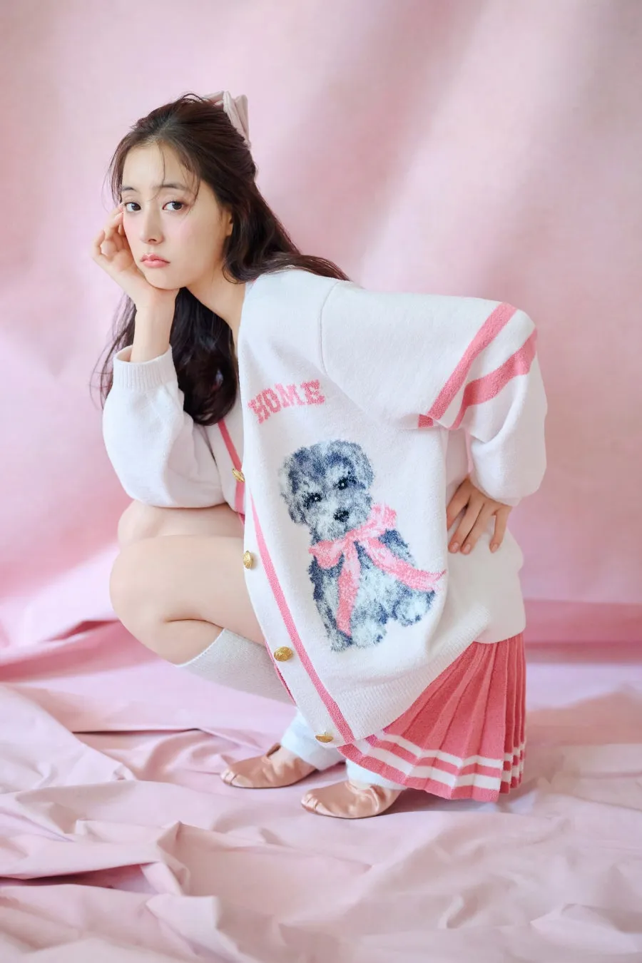 ピンク色の背景に、白いカーディガンとピンクのプリーツスカートを着た女性が座ってポーズをとっています。カーディガンには犬の絵が描かれており、「HOME」という文字が刺繍されています。女性は片手で顎に触れ、カメラを見つめています。