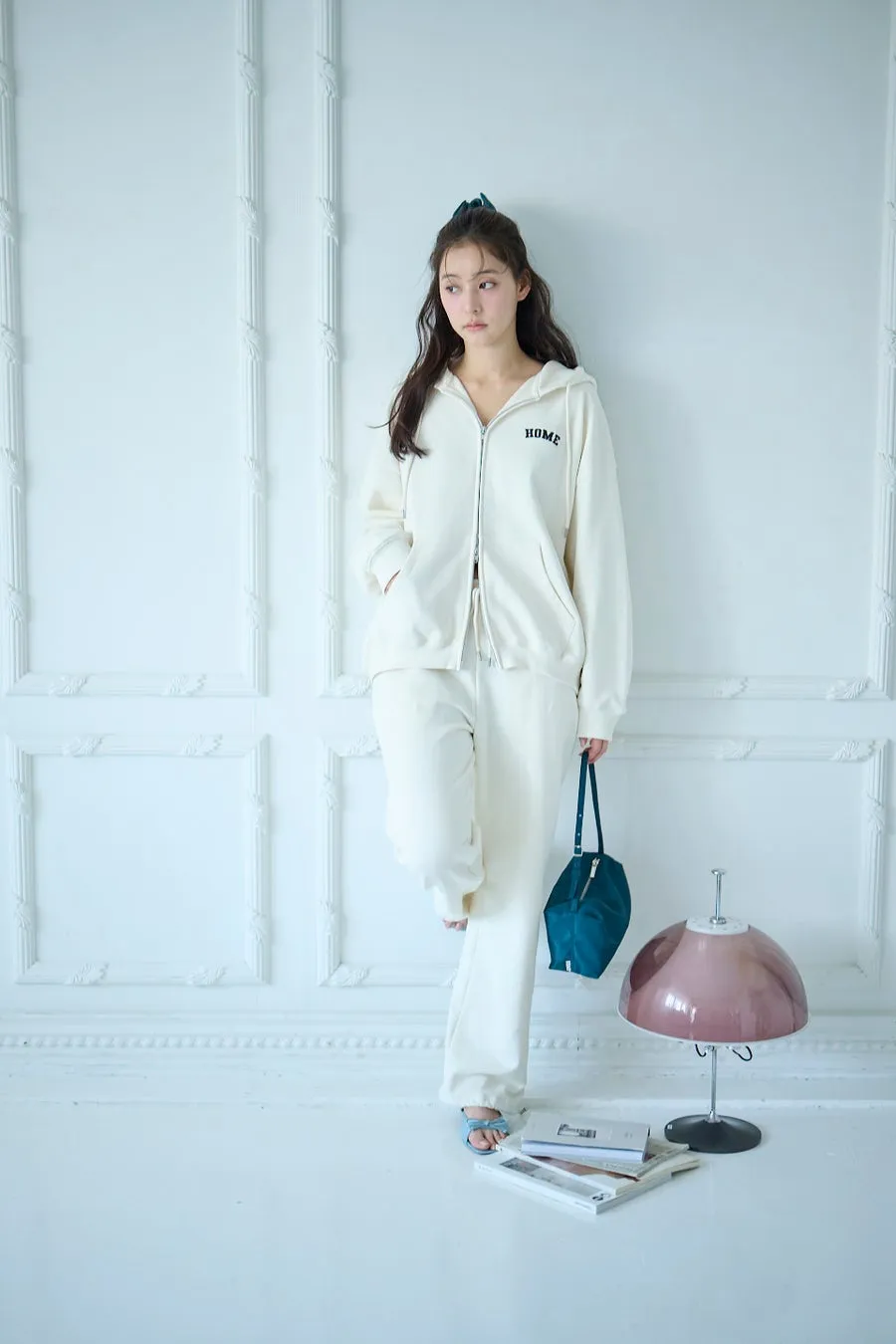 女性が白い部屋で、白いパーカーとパンツのセットアップを着て立っています。彼女は青いバッグを持っていて、足元には雑誌や本が置かれています。ピンクのランプが横にあります。