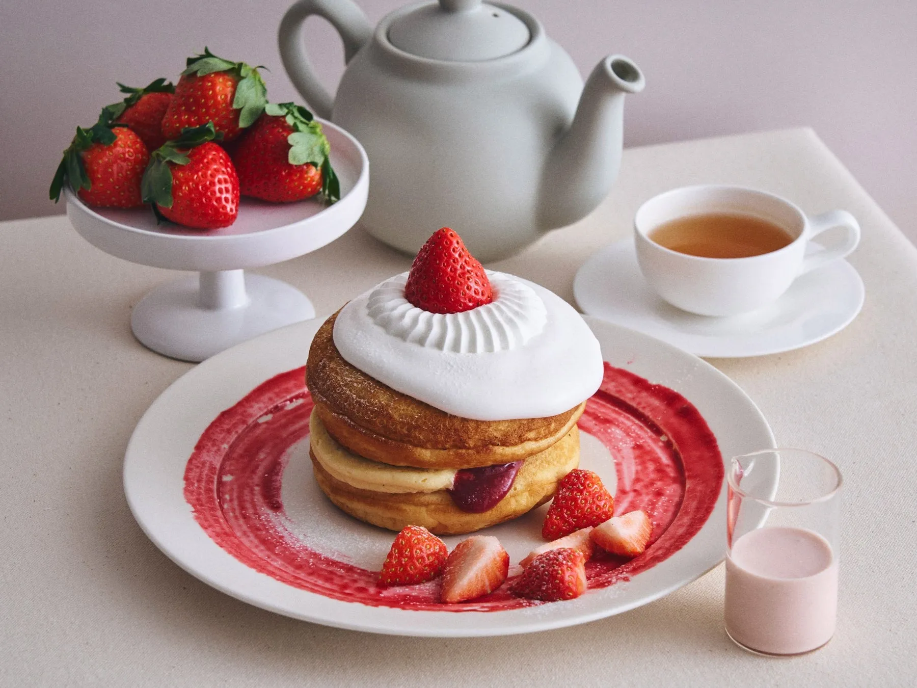 いちごのパンケーキと紅茶のセットの写真。パンケーキはホイップクリームとイチゴで飾られており、皿には赤いソースが塗られている。他に、イチゴ、紅茶、ピンク色の飲み物が写っている。