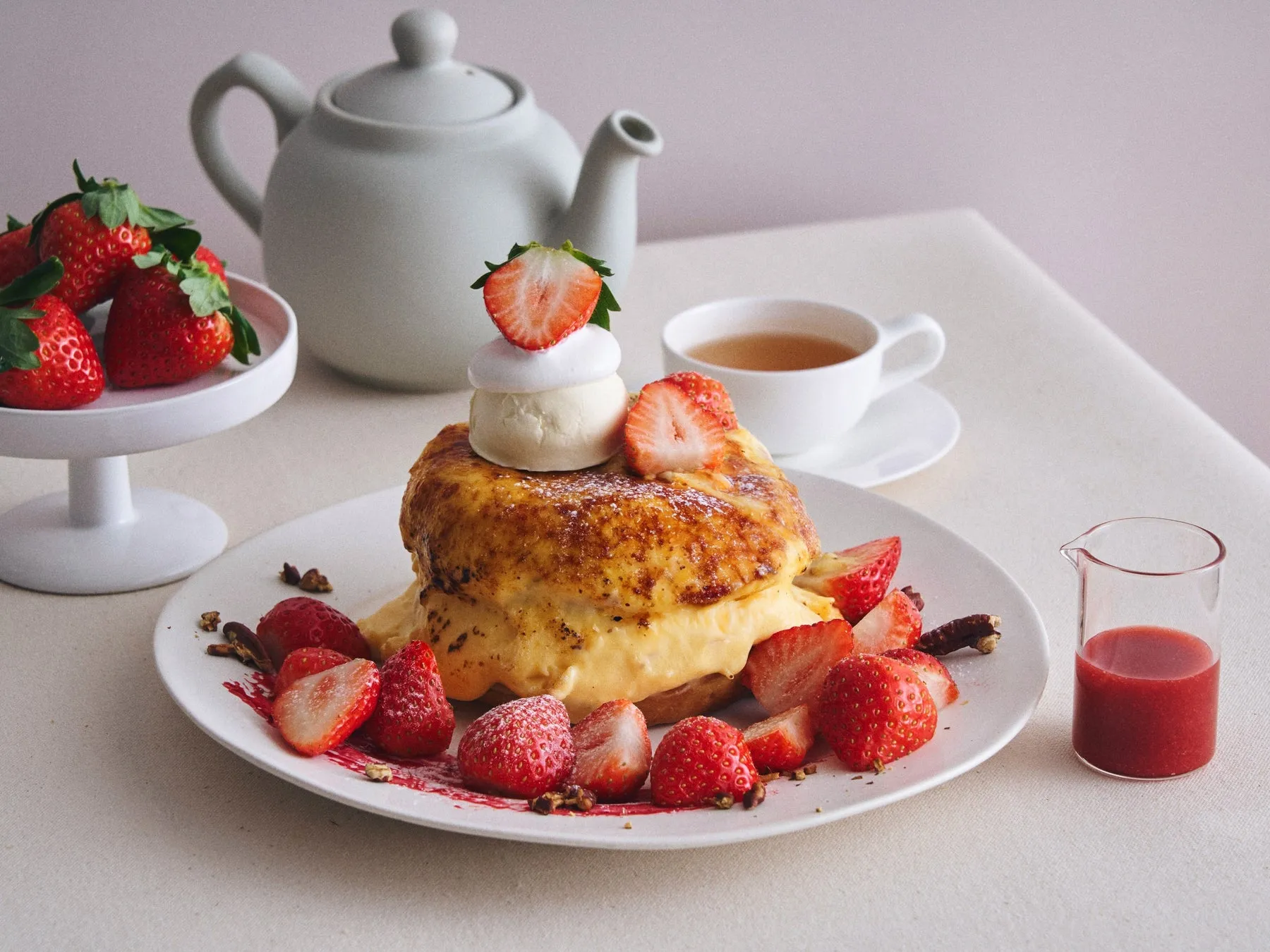 いちごが添えられたパンケーキと紅茶、ティーポットの画像。パンケーキはホイップクリームとアイスクリームで飾られており、皿にはいちご、ナッツ、ソースが添えられています。