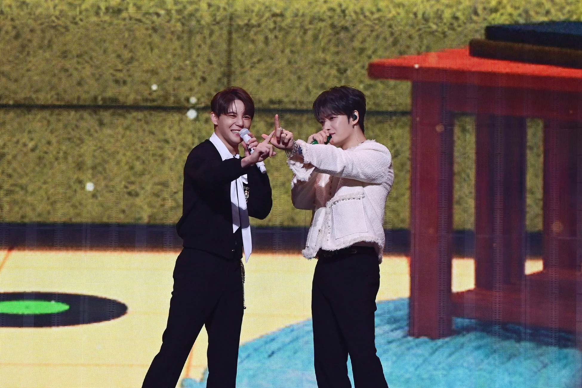 2人の男性がステージ上で歌を歌っている様子。2人ともマイクを持っており、笑顔で観客に手を振っている。背景には、赤と黄色の装飾が施されたステージセットがある。