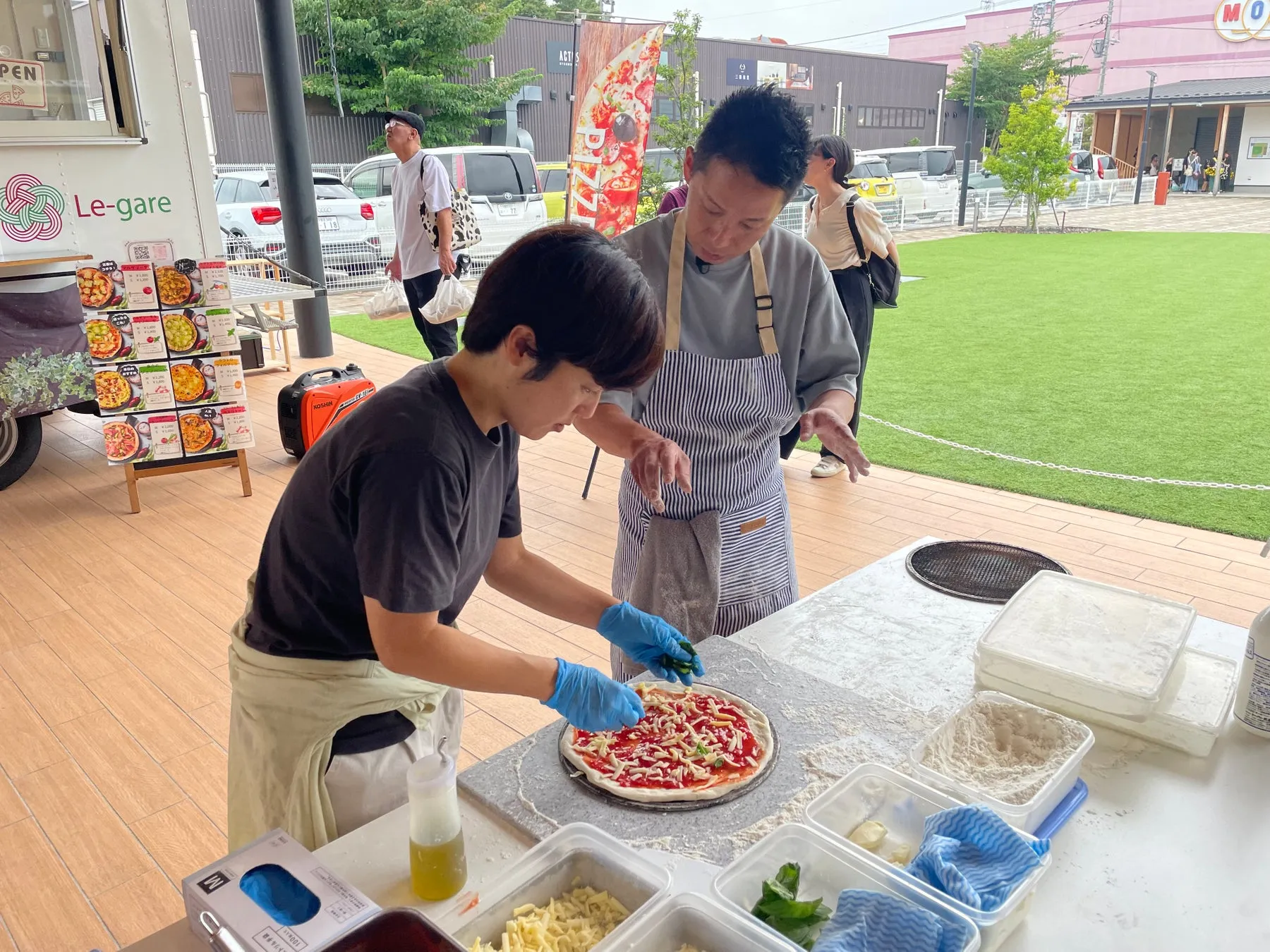 ピザを調理している二人の男性の画像。一人はピザ生地に具材を乗せ、もう一人はそれを見守っている。周囲にはピザの材料やキッチンカーがあり、屋外で調理されている様子。