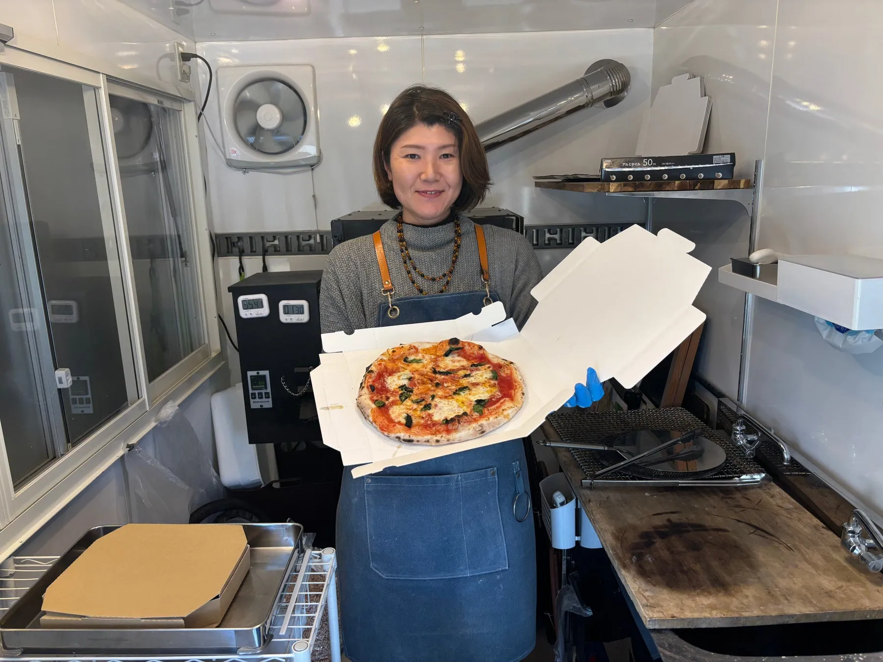 女性がピザを手に持って微笑んでいる写真。ピザは箱に入っており、彼女はエプロンを着けている。背景には調理器具や換気扇が見える。
