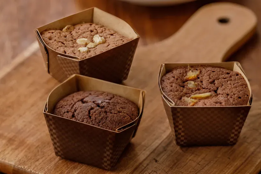 木製のまな板の上に、3つのチョコレートケーキが紙製のカップに入って配置されています。ケーキはそれぞれ異なるトッピングが施されており、手前と奥のケーキにはナッツが、右側のケーキには柑橘系の果物がトッピングされています。