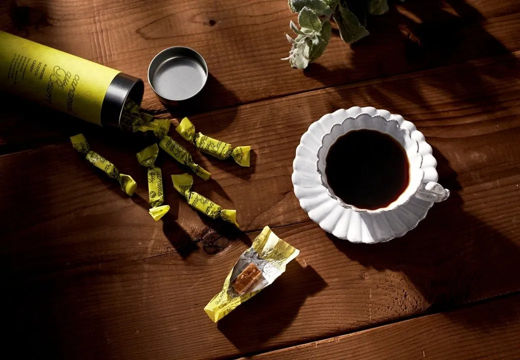 木製のテーブルの上に、キャラメルとコーヒーカップが置かれています。キャラメルは黄色い包装紙に包まれており、一部は包装が解かれています。コーヒーカップは白く、コーヒーが注がれています。全体的に、温かみのある雰囲気の写真です。