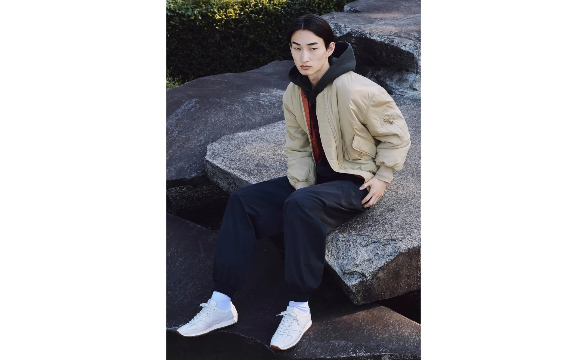 岩の上に座っている男性。彼はベージュのボンバージャケット、黒いパーカー、黒いパンツ、白い靴を履いています。