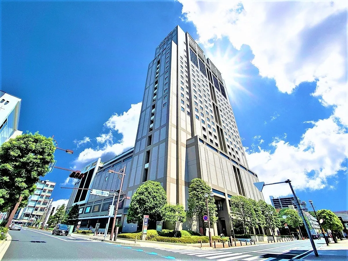 青空の下、高層ビルの写真。街路樹や車、歩行者用信号機、横断歩道が見える。