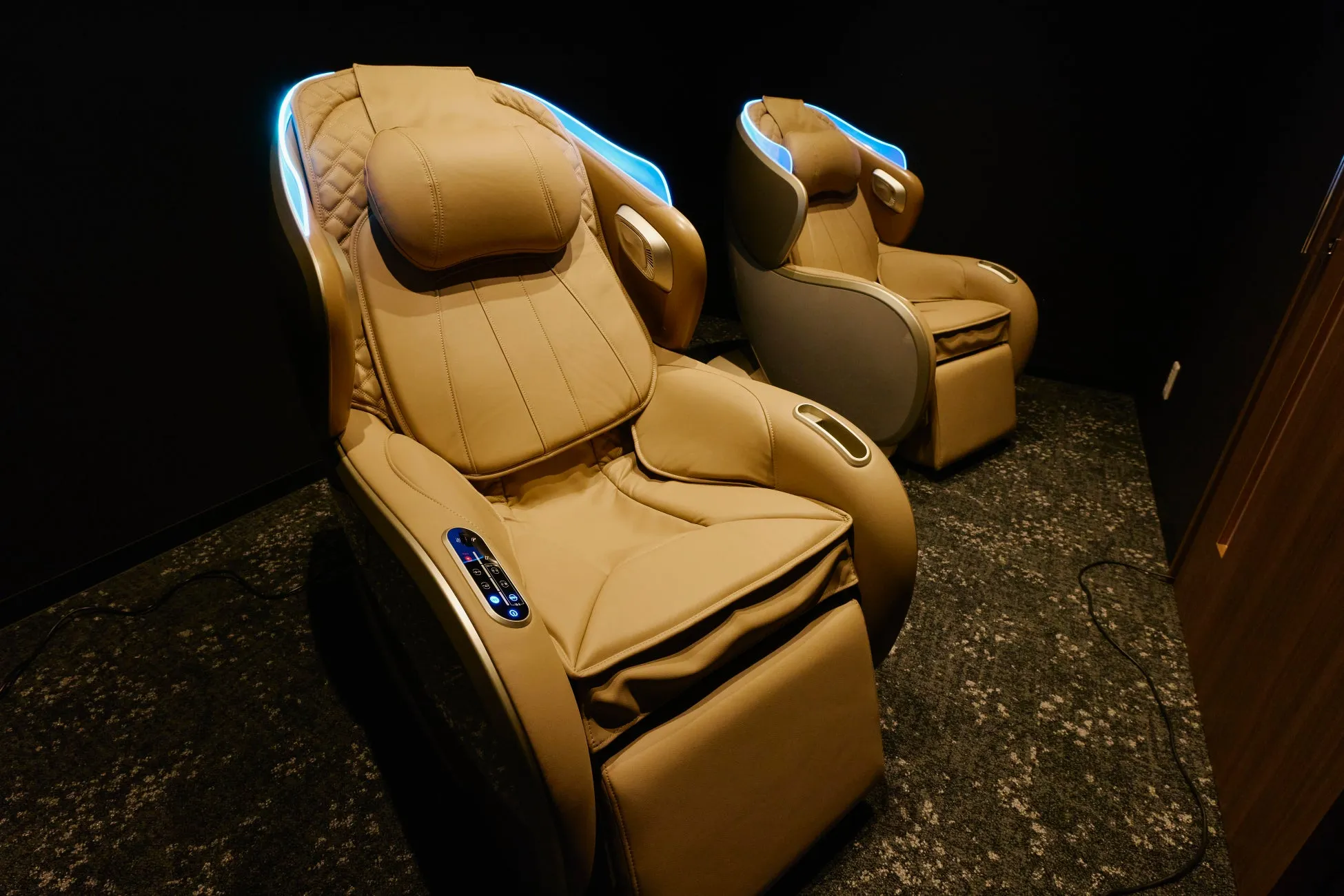 2つのマッサージチェアが並んだ写真です。チェアはベージュ色で、青色のライトが装飾されています。背景は黒色で、床は模様のあるカーペットです。