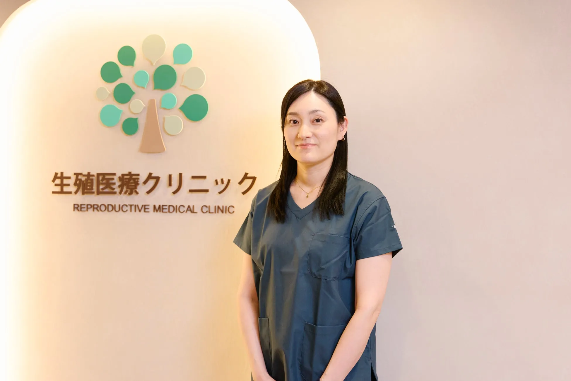 女性がクリニックの受付に立っている写真。背景にはクリニックのロゴと看板が写っています。