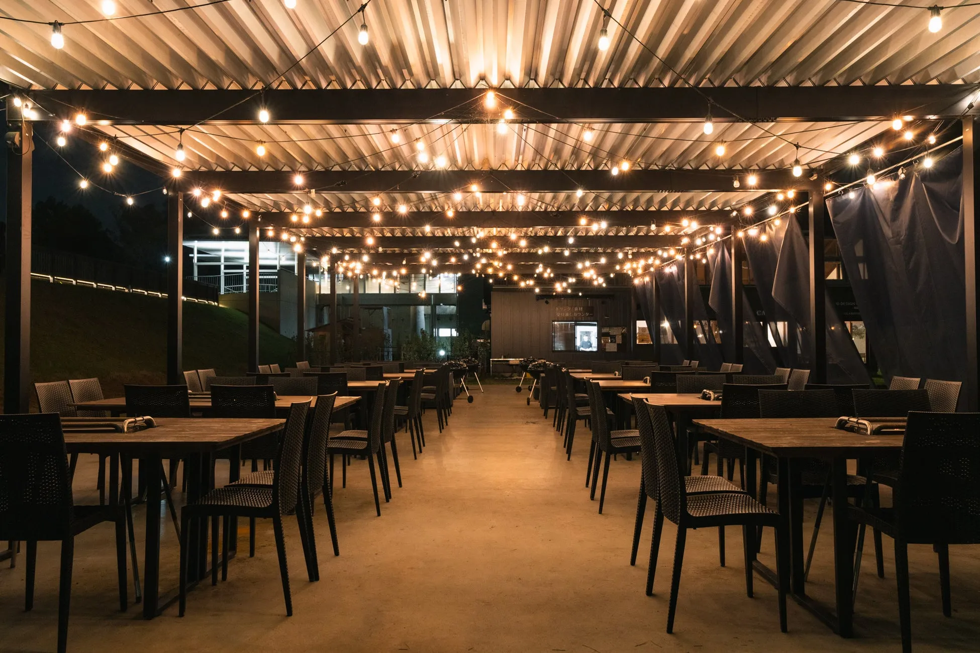 電球が飾られた屋外のレストランの夜景。テーブルと椅子が並び、奥には建物が見える。