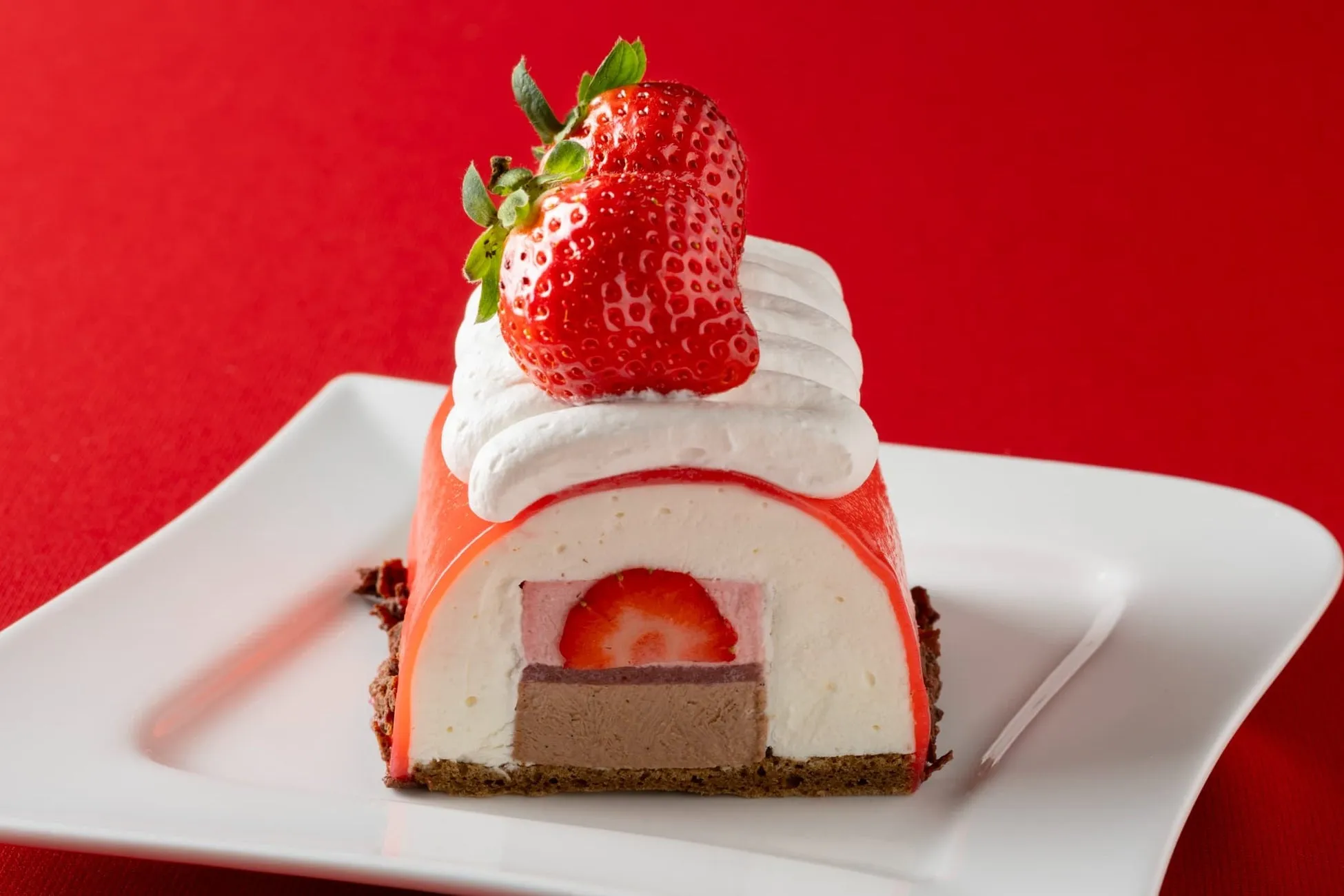 いちごがトッピングされた、断面が美しいケーキの写真です。赤色の背景に、白の皿に載せられています。