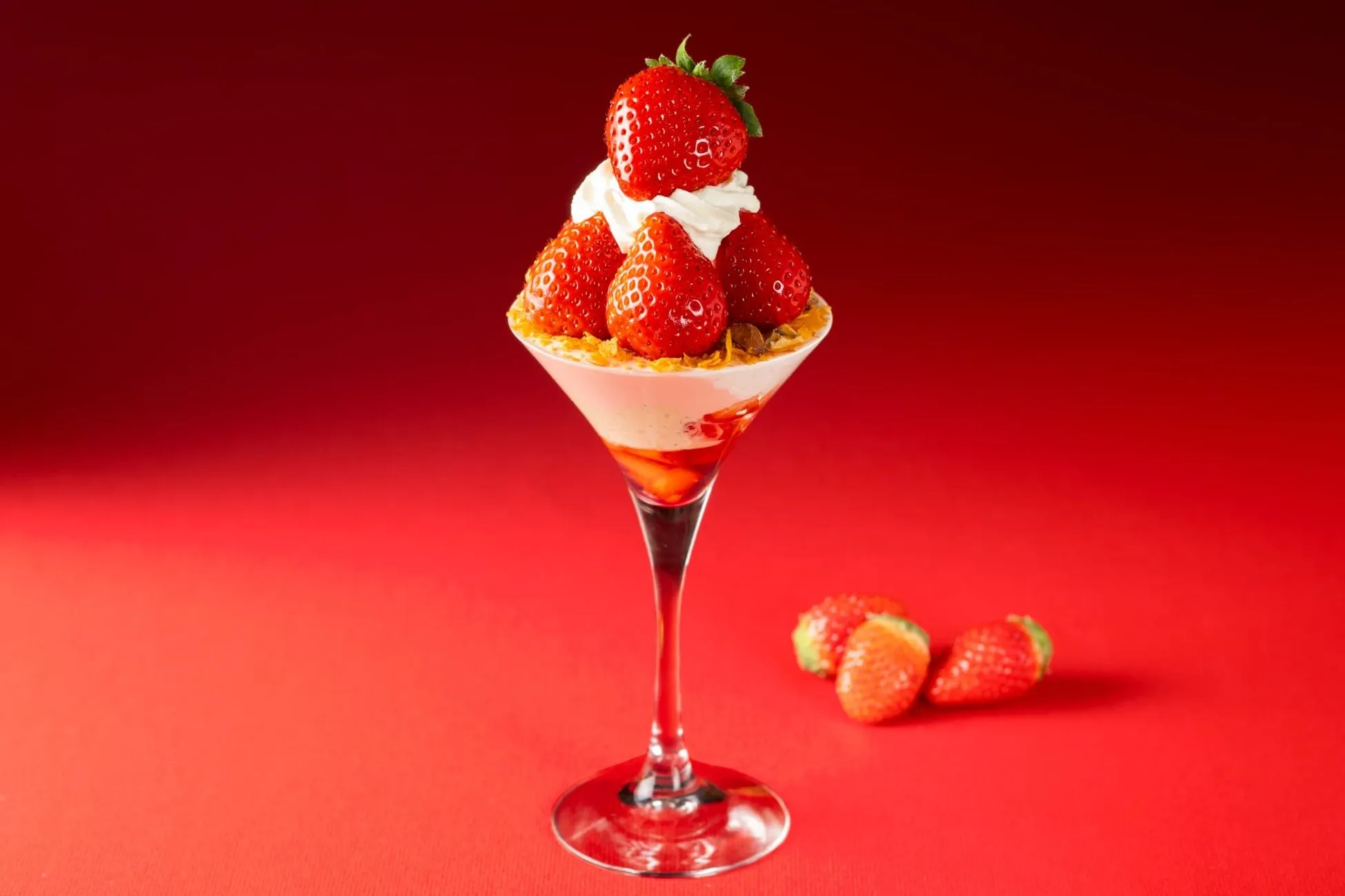 赤い背景に、イチゴをトッピングしたパフェがグラスに入って置かれています。グラスの横にはイチゴがいくつか置かれています。