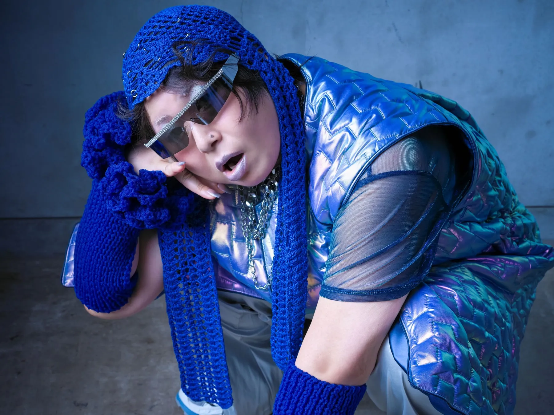 青色のニット帽、サングラス、青色のベストを着て、片手で顔を支えている人物のクローズアップ写真。背景は青色の壁。