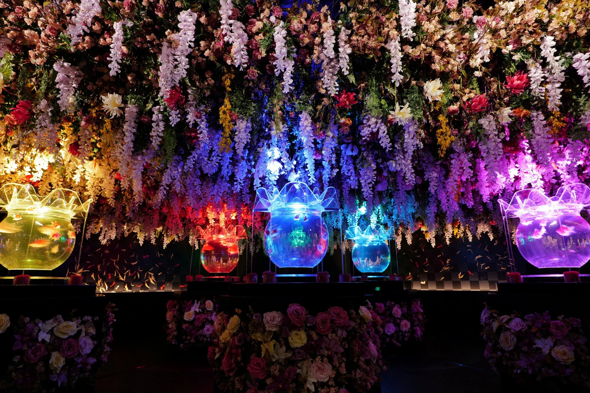 色とりどりの花々が咲き乱れる空間に、金魚鉢が並んでいる様子。金魚鉢はそれぞれ異なる色にライトアップされ、幻想的な雰囲気を醸し出している。天井からは花々が垂れ下がり、華やかさを添えている。