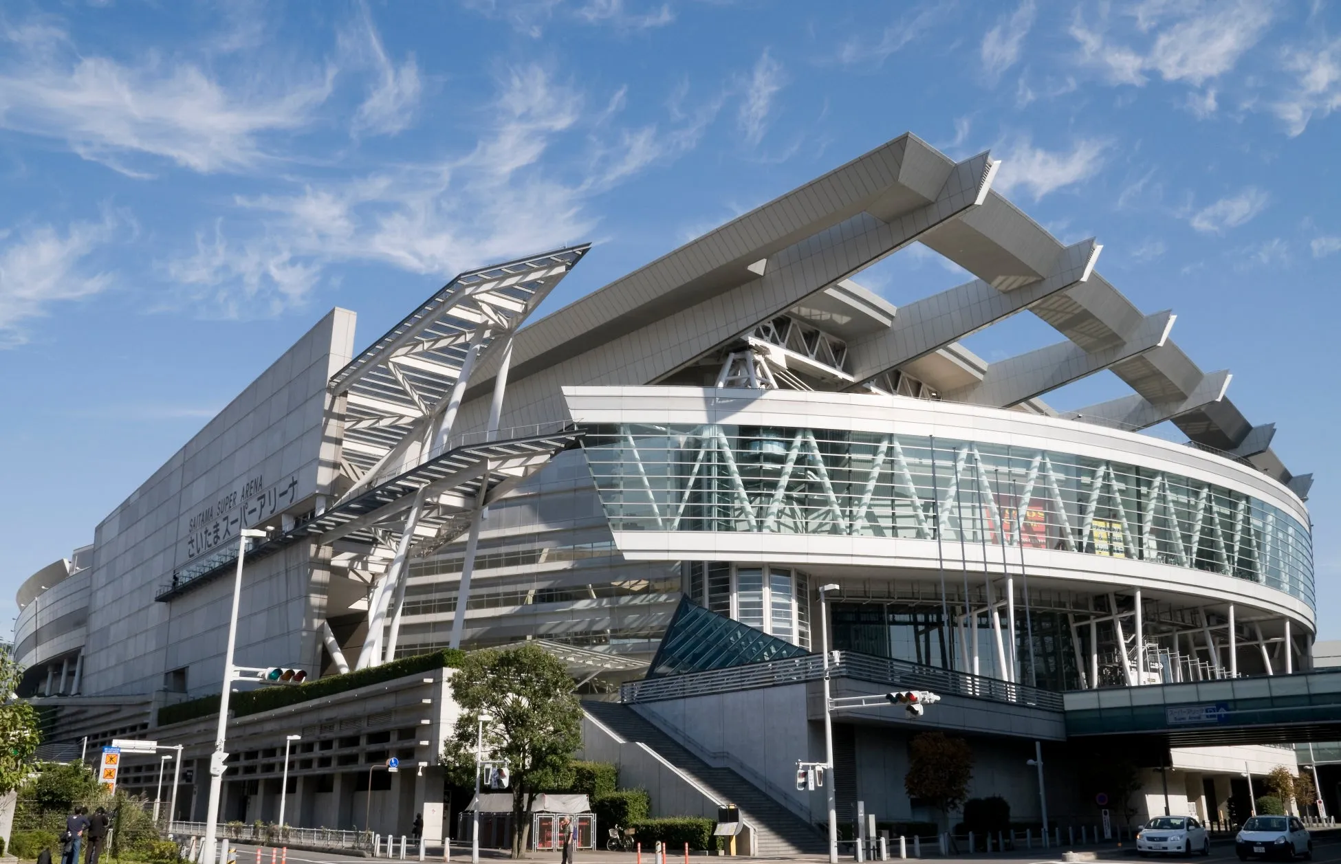青空の下、さいたまスーパーアリーナの全景を捉えた写真。建物の外観は近代的なデザインで、ガラス張りの壁面と特徴的な屋根構造が印象的。周辺には街路樹や歩道があり、都市的な風景が広がっている。