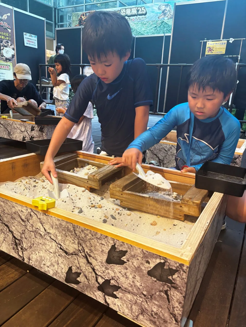 子供たちが砂場で化石の発掘体験をしている様子。砂場には恐竜の足跡が描かれており、スコップを使って砂を掘り起こしている。周りには、他の子供や大人もおり、イベントの賑やかさが伝わってくる。