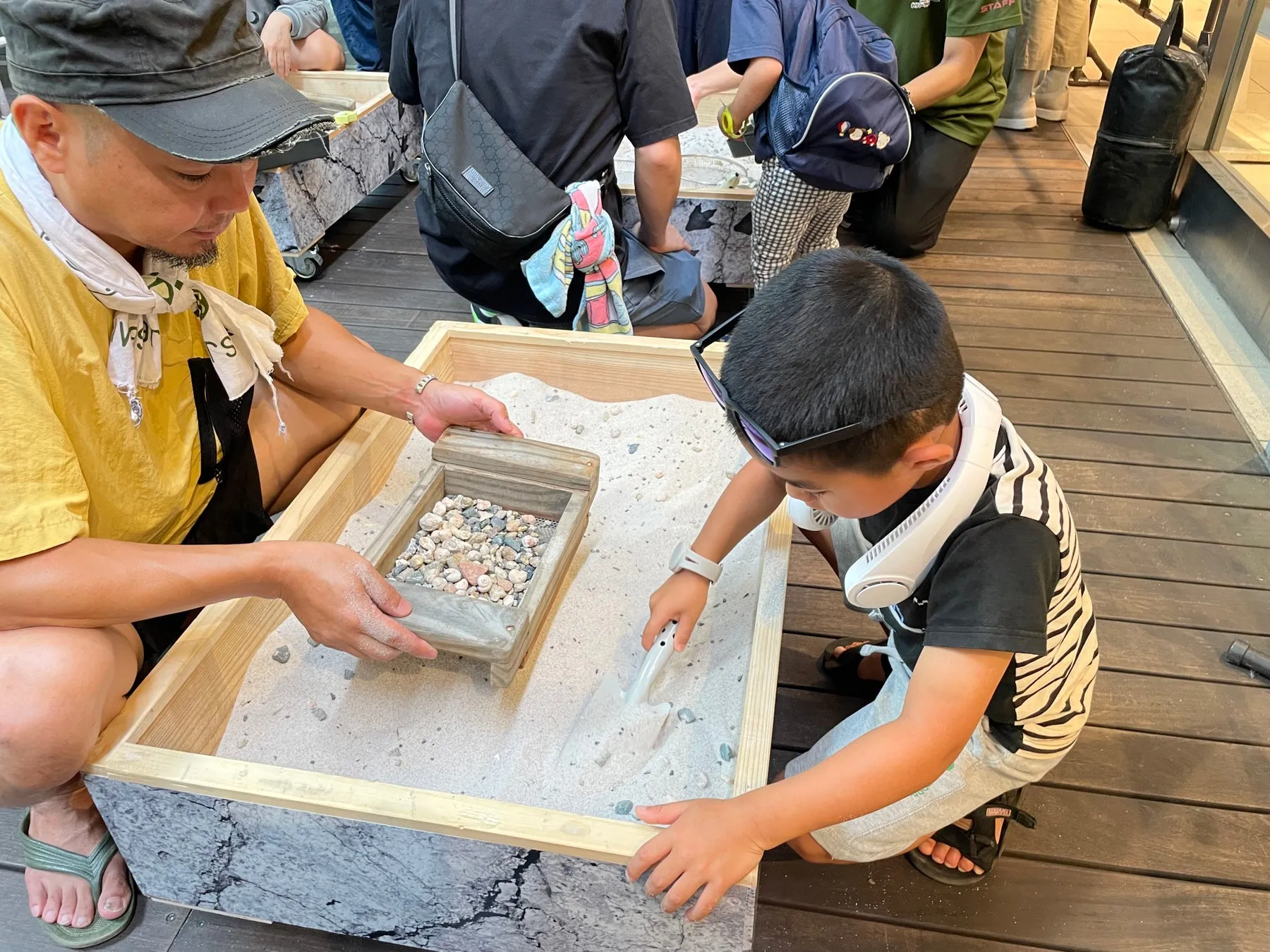 砂場のような場所で、子供と大人が一緒に遊んでいる様子を捉えた写真です。子供は砂を掘り、大人は砂が入った木製の箱を持っています。周りには他の人もいて、何かを探しているようです。