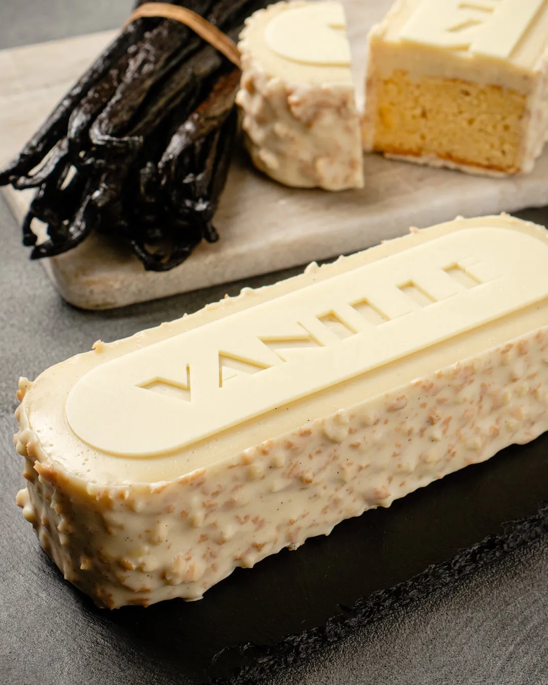 バニラビーンズとバニラ風味のケーキの写真。ケーキは長方形で、表面にVANILLAの文字が刻まれている。バニラビーンズはケーキの横に置かれている。