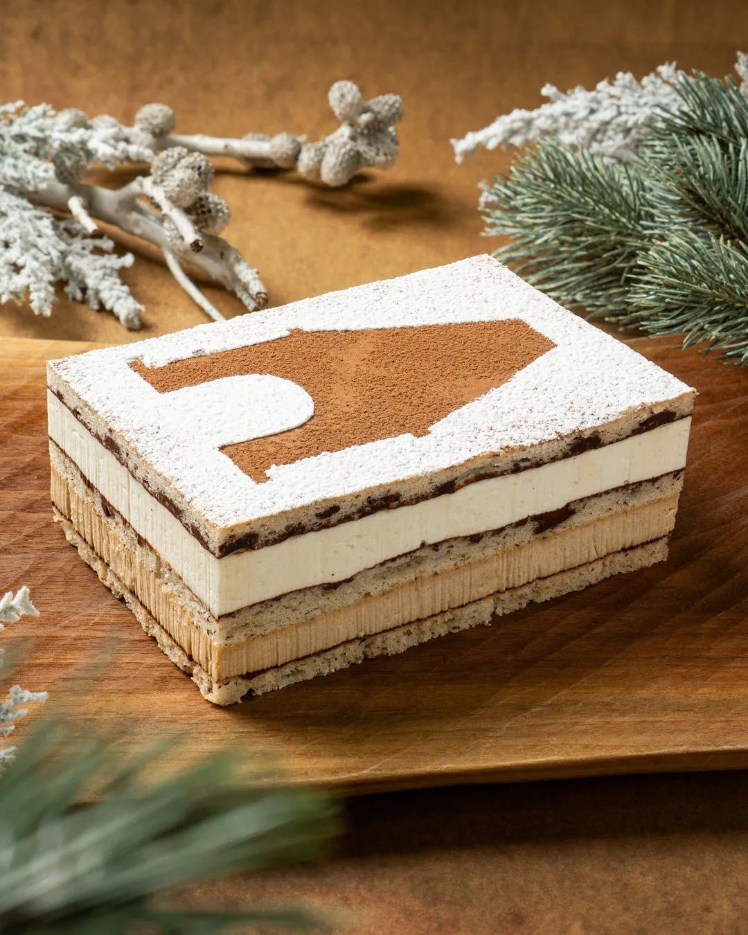 木製の皿の上に、ココアパウダーでデコレーションされた長方形のケーキが置かれています。ケーキの周りには、クリスマスの装飾が施されています。