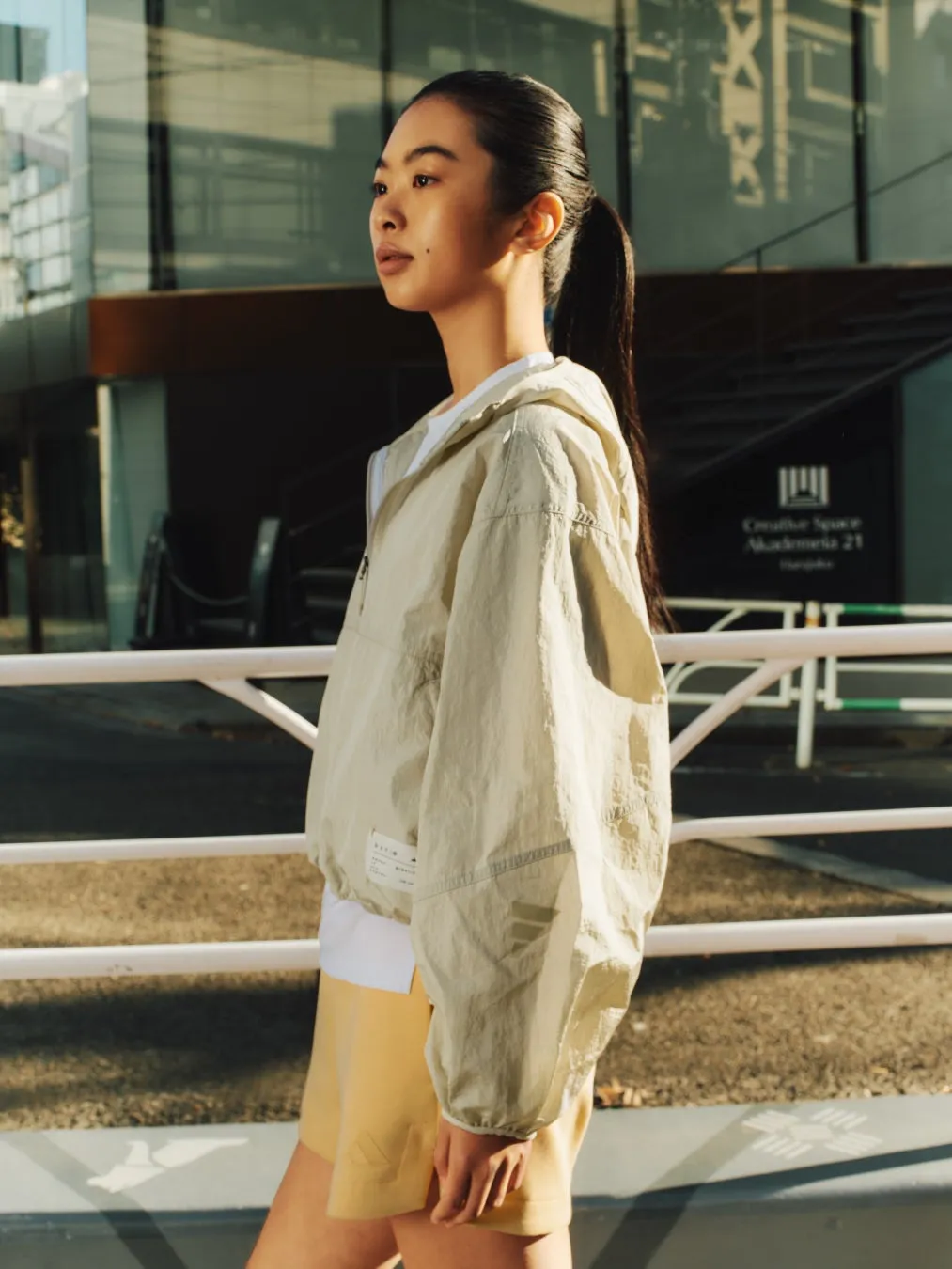 女性が屋外でポーズをとっている写真。彼女はベージュのジャケット、白いシャツ、黄色のショーツを着ています。彼女はポニーテールをしており、都市の背景に立っています。