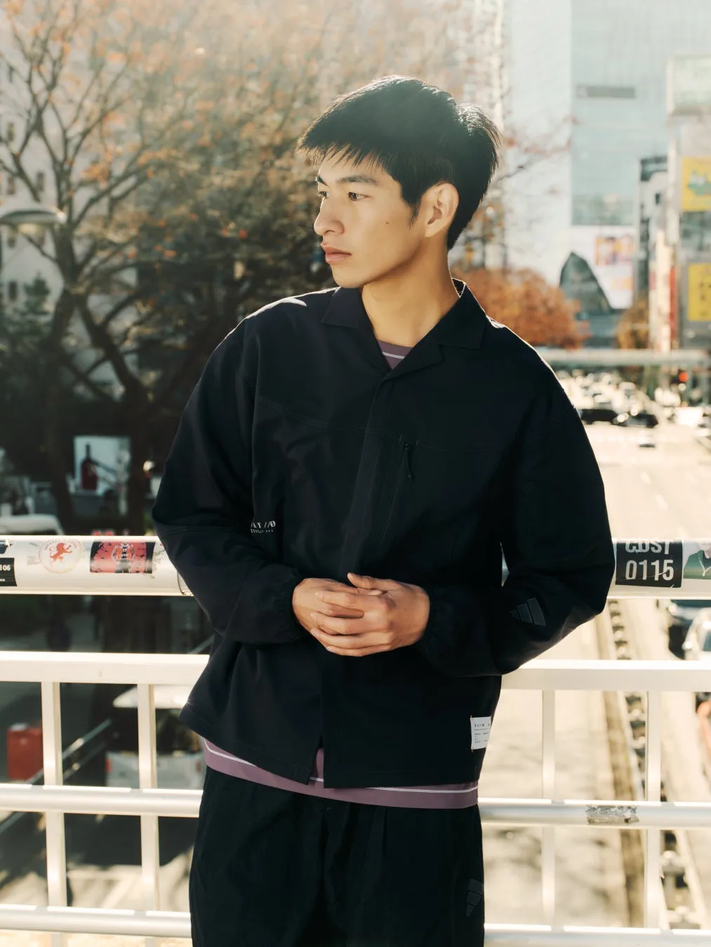 橋の上でポーズをとる男性。彼は黒いジャケットとズボンを着ており、都市の風景を背景にしています。