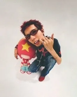 サングラスをかけた男性が、赤い人形を抱きかかえて立っている写真。男性は、赤い髪、黒いTシャツ、ジーンズを着用している。人形は、赤いボディに黄色の星のマークがついている。