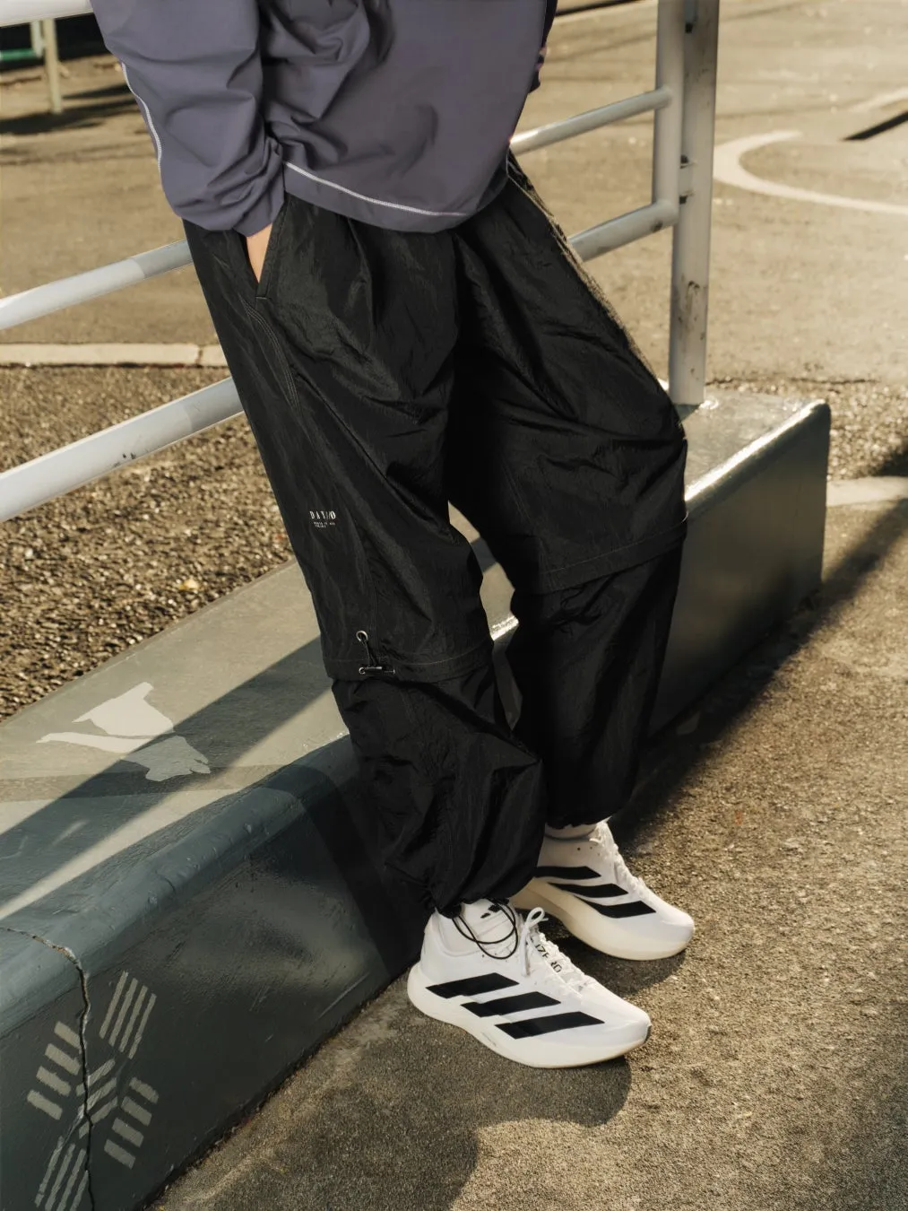 黒いパンツと白いスニーカーを履いた人物の画像。アスファルトの上で、手すりに寄りかかっている。
