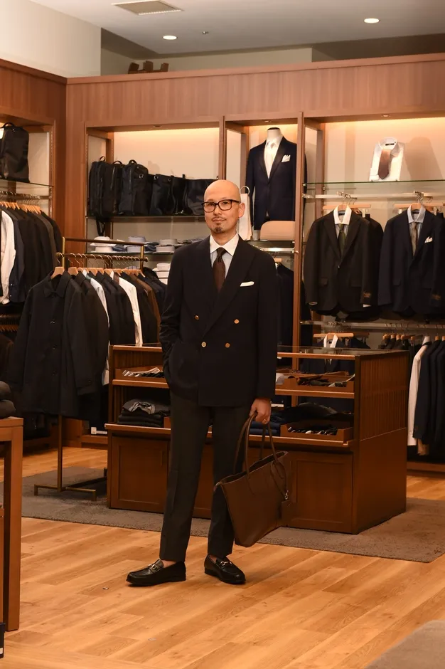 男性がスーツを着て、革のバッグを持ち、店の前に立っている様子。