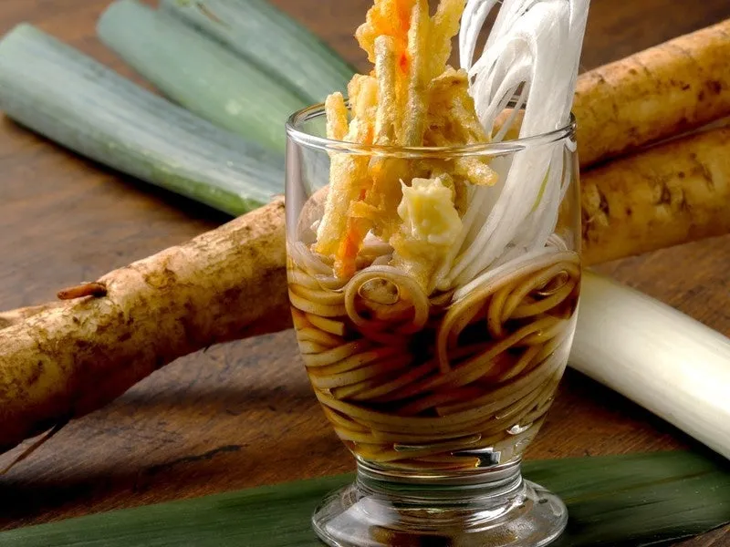 ガラスの器に入った蕎麦。天ぷら、ネギ、蕎麦、出汁、ゴボウが添えられている。周りにはゴボウ、ネギ、葉が置かれている。