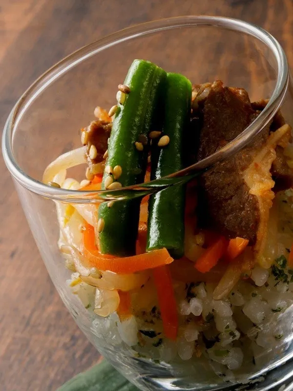 透明なグラスに入った料理の写真。ご飯の上に、インゲン、人参、肉、ゴマなどが盛り付けられている。