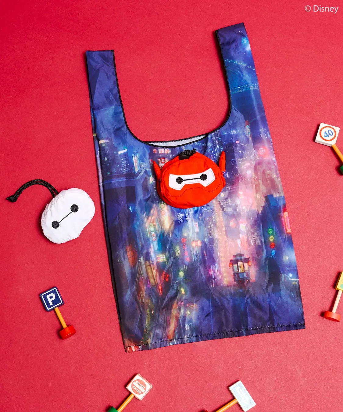 赤い背景に、ベイマックスの折り畳みエコバッグ、ベイマックスのぬいぐるみ、ミニチュアの標識が配置された写真