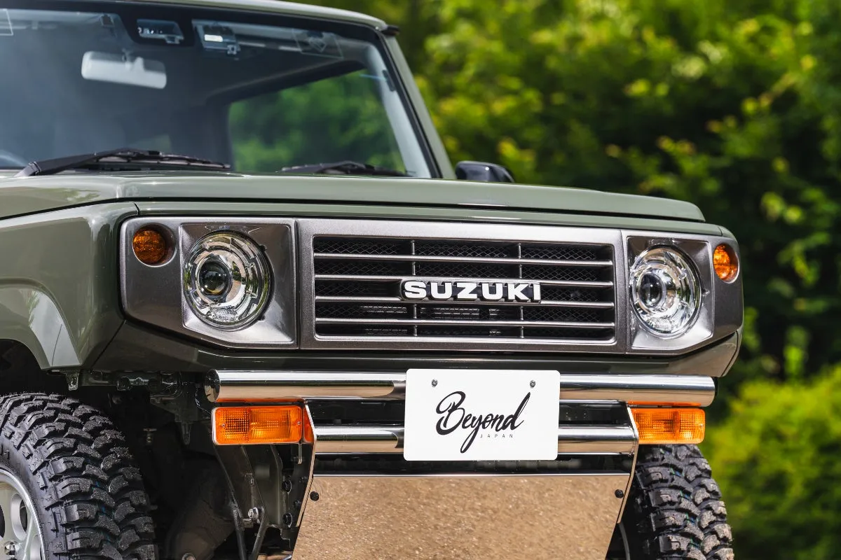 スズキのジムニーのフロント部分を捉えた写真。グリルには「SUZUKI」のロゴがあり、ナンバープレートには「Beyond JAPAN」と書かれています。背景には緑の木々が写っています。