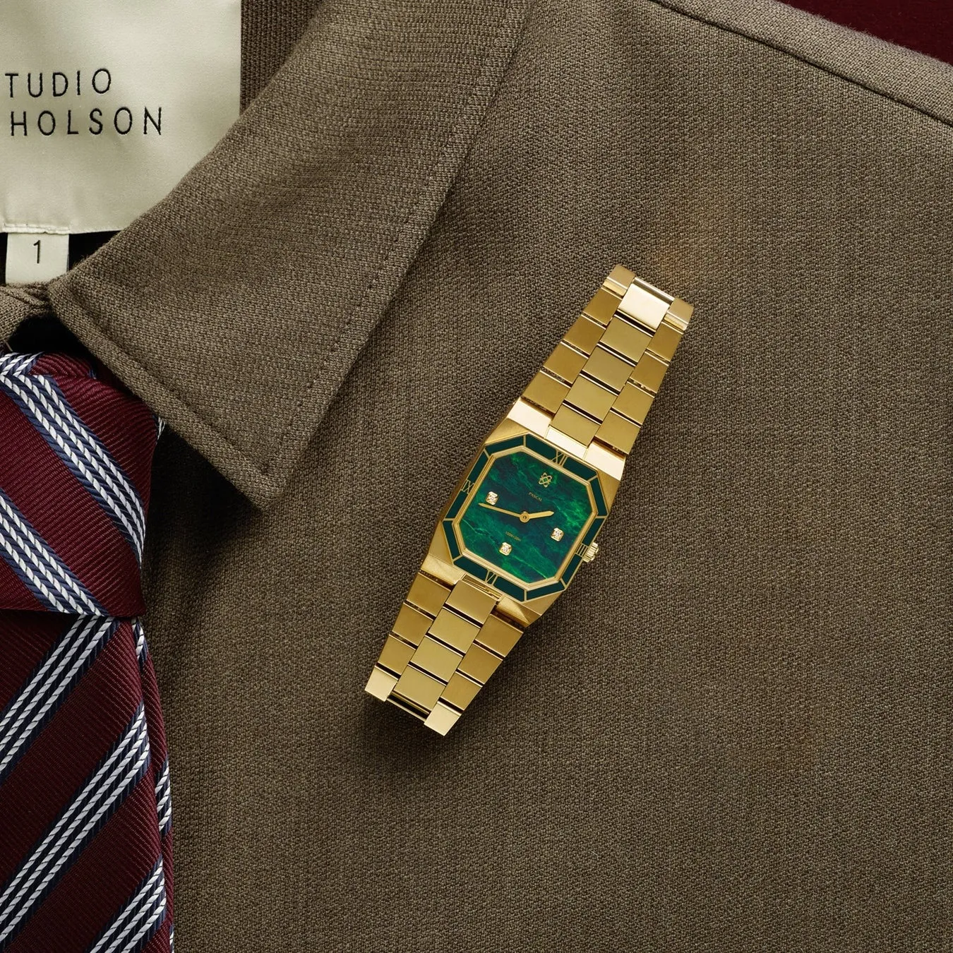 茶色のスーツの上に、緑色の文字盤と金色のバンドを備えた金色の時計が置かれています。また、赤いストライプのネクタイも見えます。
