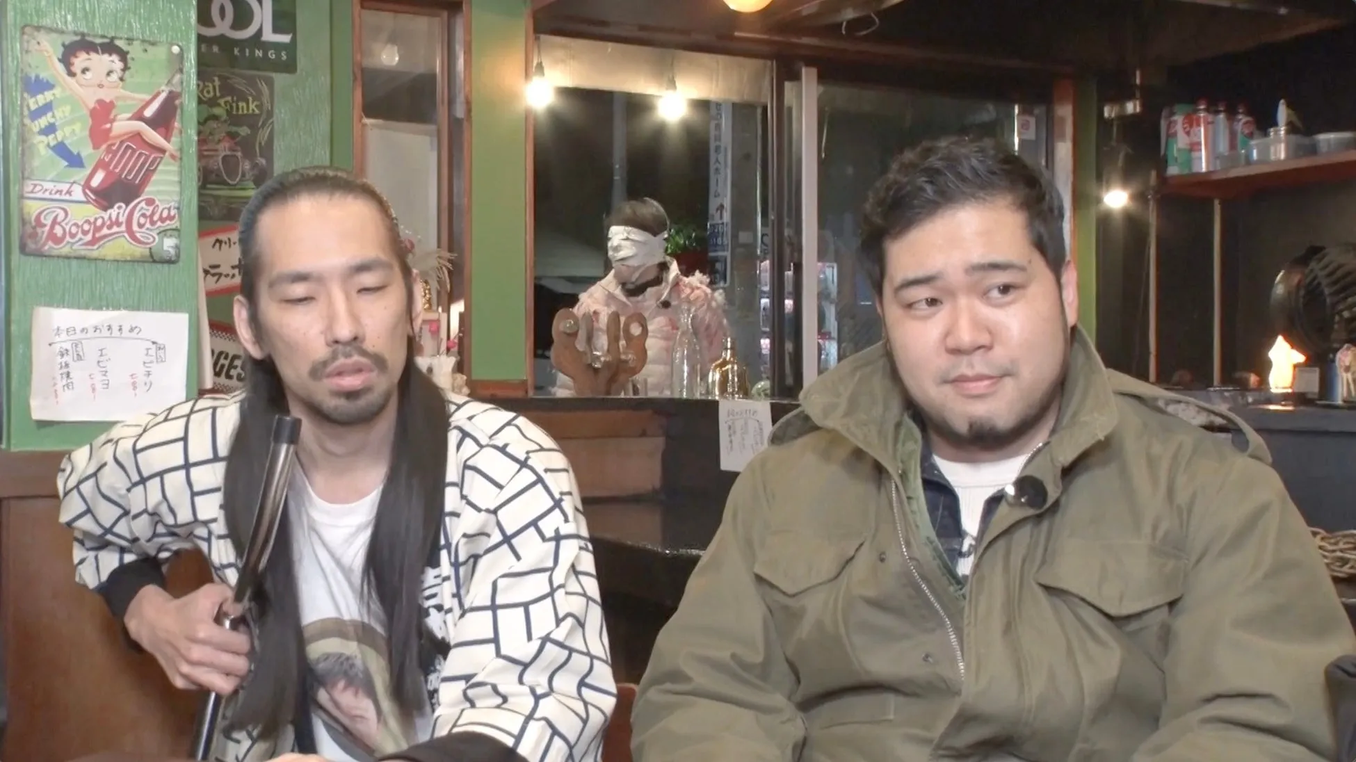 2人の男性がバーのような店内で話している様子。背景にはベティ・ブープのポスターや、目隠しをした人物が立っているのが見える。