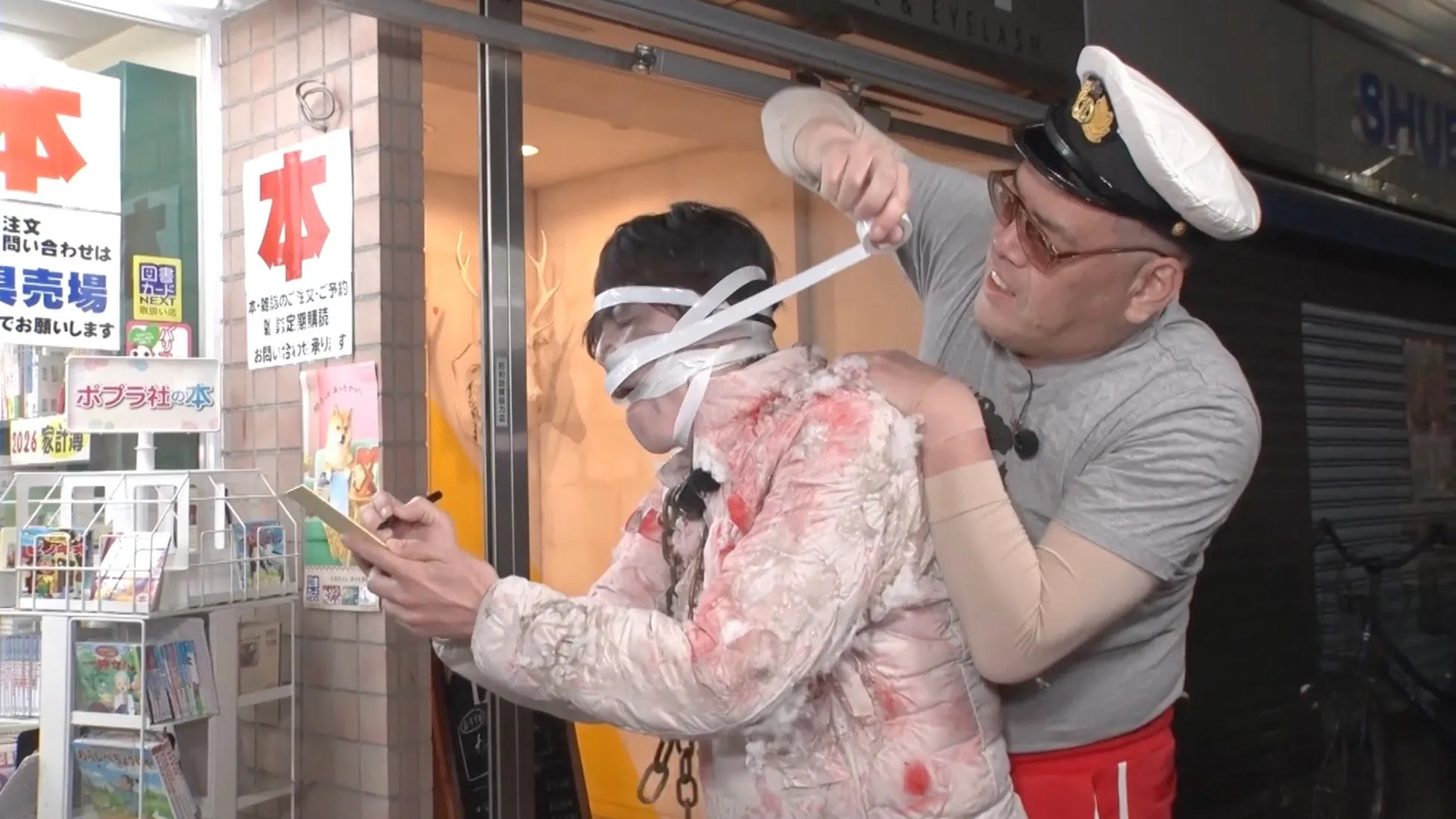 男性2人が店内で撮影している様子。一人の男性は目隠しされ、血のようなものが付着した服を着ており、もう一人の男性は彼を拘束しようとしているように見える。