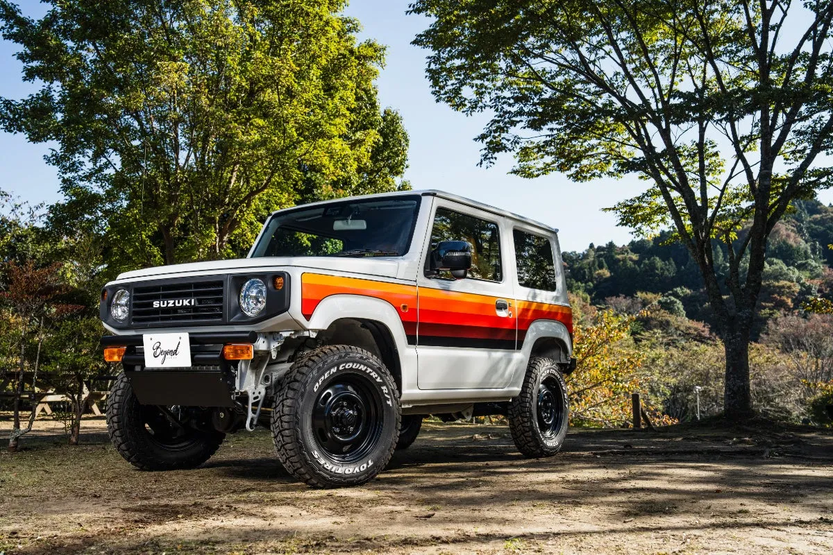 白いスズキジムニーが草地の上に駐車されています。車体にはオレンジ、赤、黒のストライプが施されています。背景には緑の木々や山々が見えます。