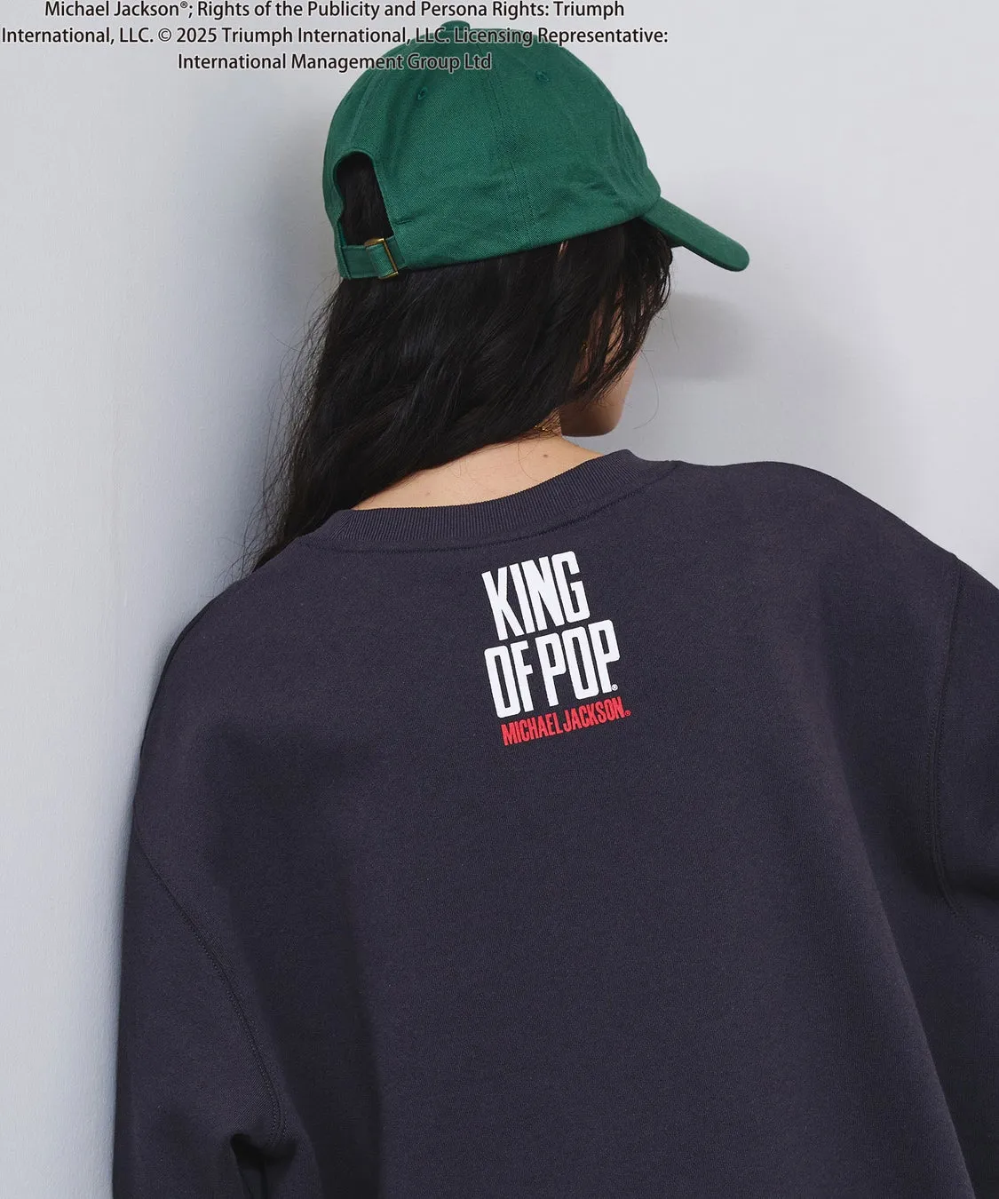 後ろ姿の女性が、マイケル・ジャクソンの「KING OF POP」と書かれたトレーナーを着て、緑色の帽子をかぶっている画像です。