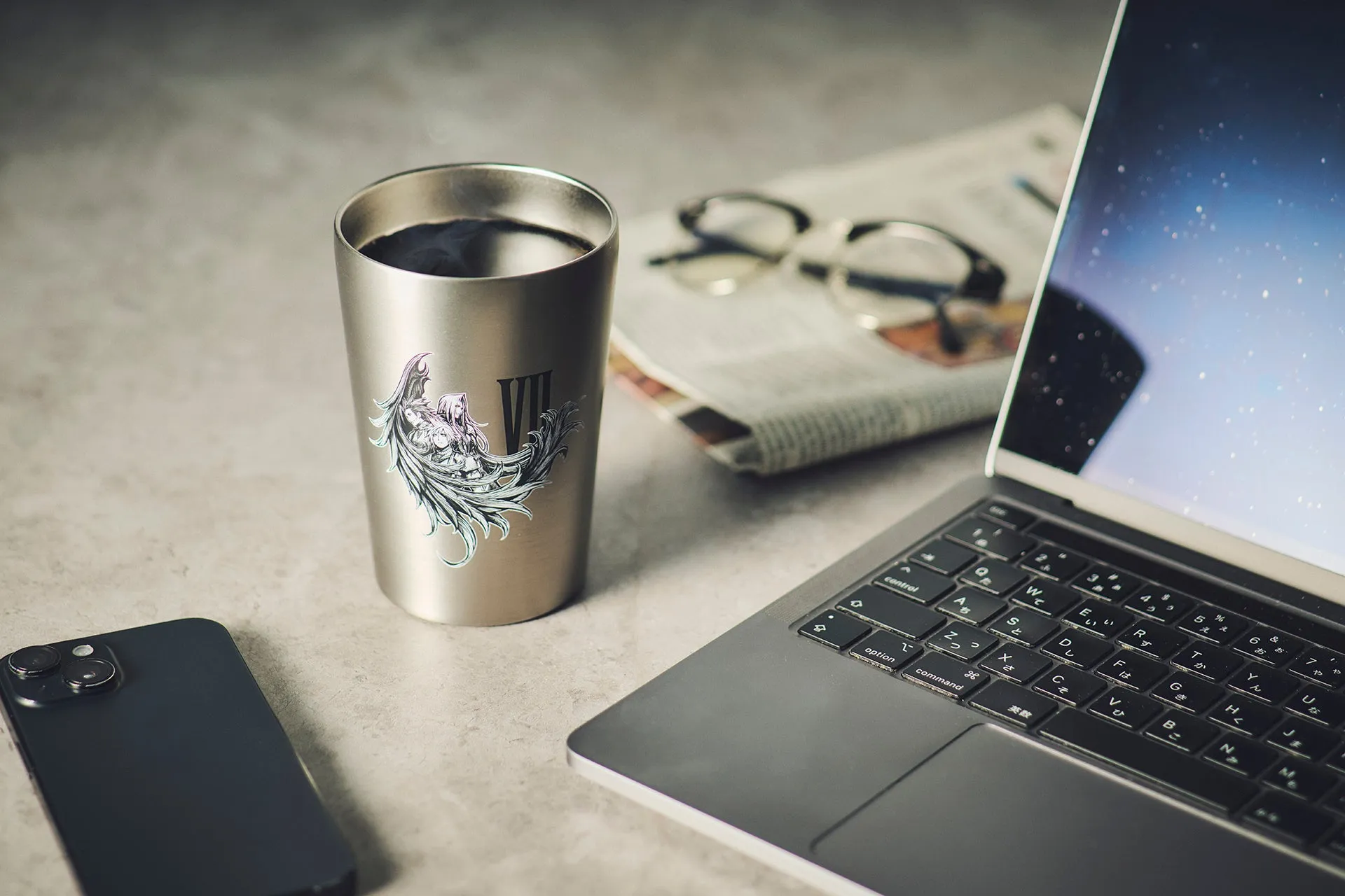 Final Fantasy VIIのキャラクターが描かれたタンブラーと、MacBook、スマートフォン、メガネ、新聞が置かれたデスクの写真