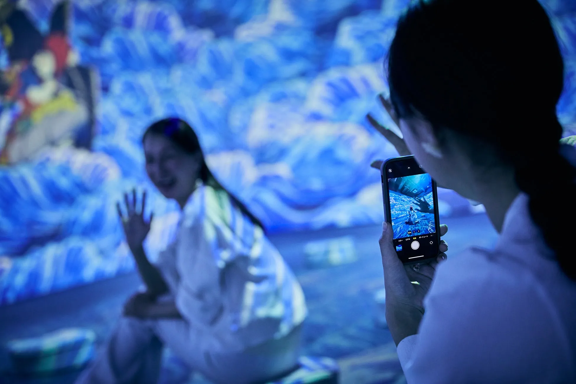 女性がスマートフォンで写真を撮っている様子。背景には青色の波模様の展示があり、もう一人の女性が手を振っている。