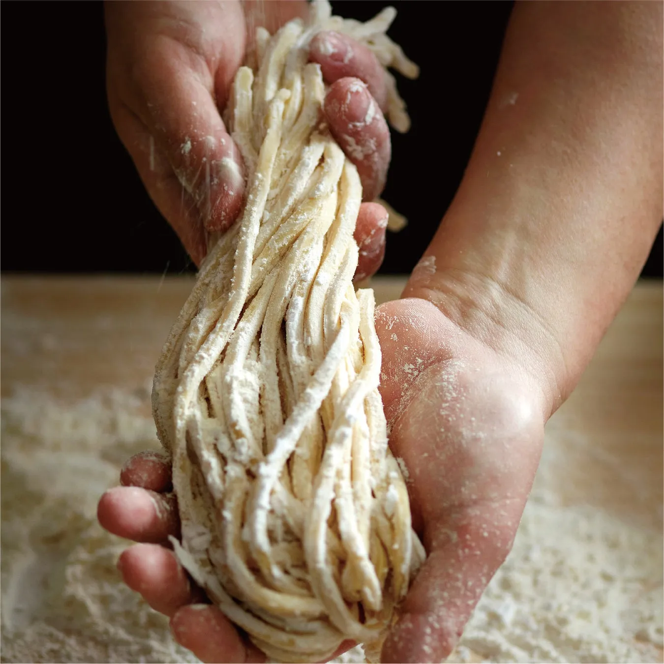 粉まみれの手が麺を掴んでいる様子を捉えたクローズアップ写真。麺は手打ち麺のようで、小麦粉がまぶされている。背景は暗く、麺と手の質感が際立っている。