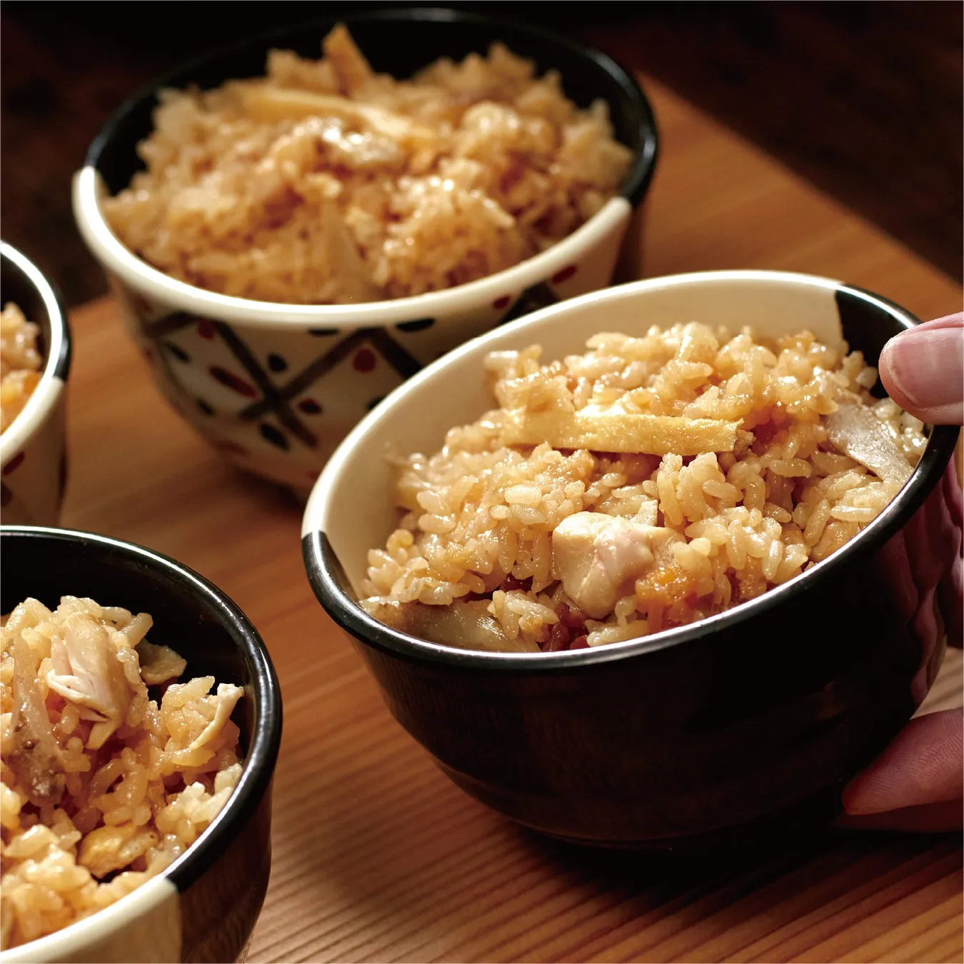 鶏肉や野菜が入った炊き込みご飯が、器に盛り付けられている様子を捉えた写真です。器は黒と白を基調としたデザインで、木製のテーブルの上に置かれています。手で器が持たれており、食事の場面を切り取ったような写真です。