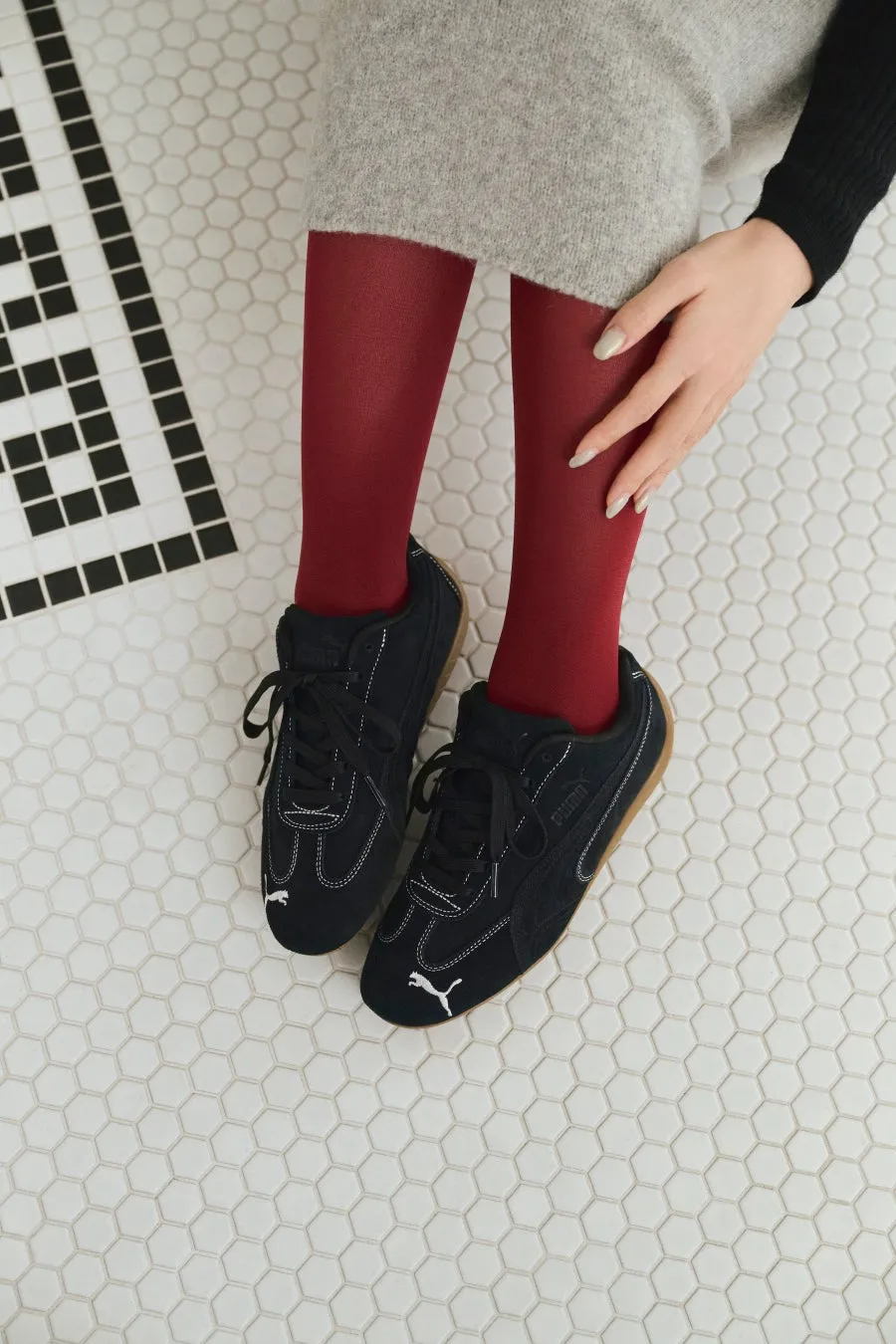 黒いスニーカーと赤いタイツを履いた女性。床は六角形のタイルで、片方の手はスカートの上に置かれています。