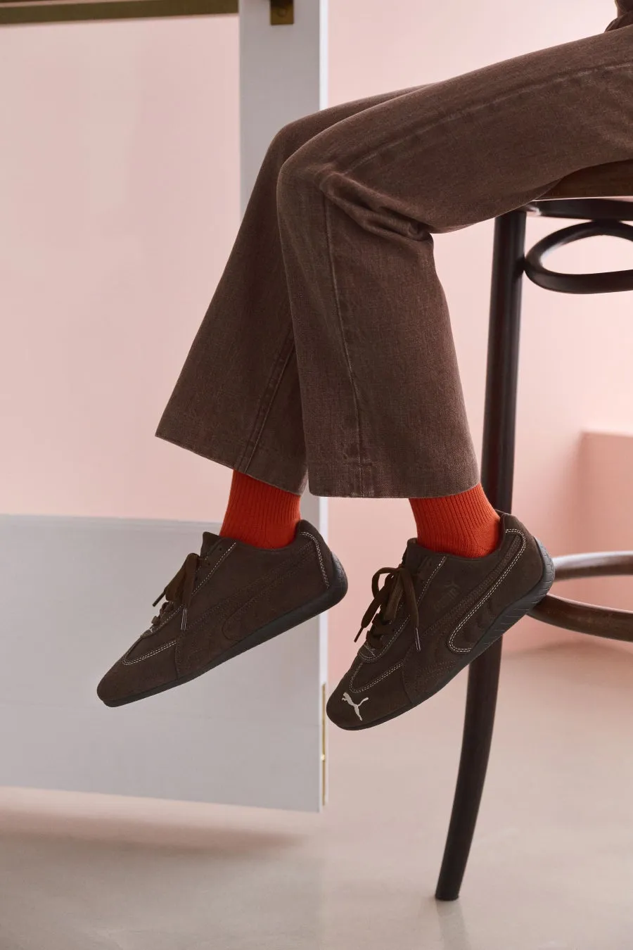 茶色のパンツとオレンジ色の靴下を履いた人が、茶色の Puma のスニーカーを履いている写真です。背景はピンク色の壁と白いドアです。