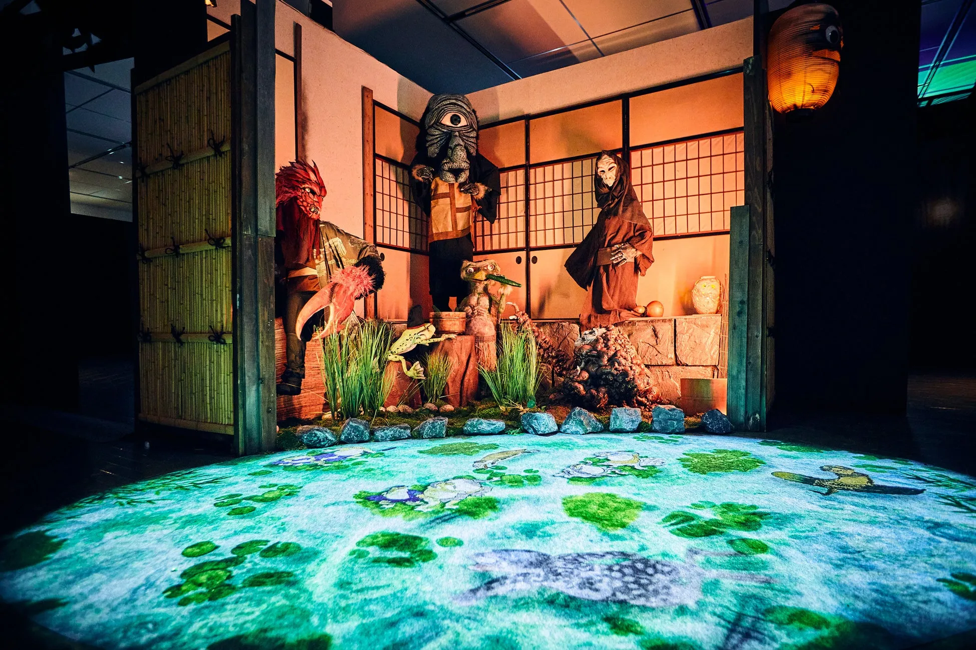 日本の妖怪をモチーフにした展示空間の様子。妖怪のマスクをつけた人物が配置され、床には水面を模したプロジェクションマッピングが施されている。